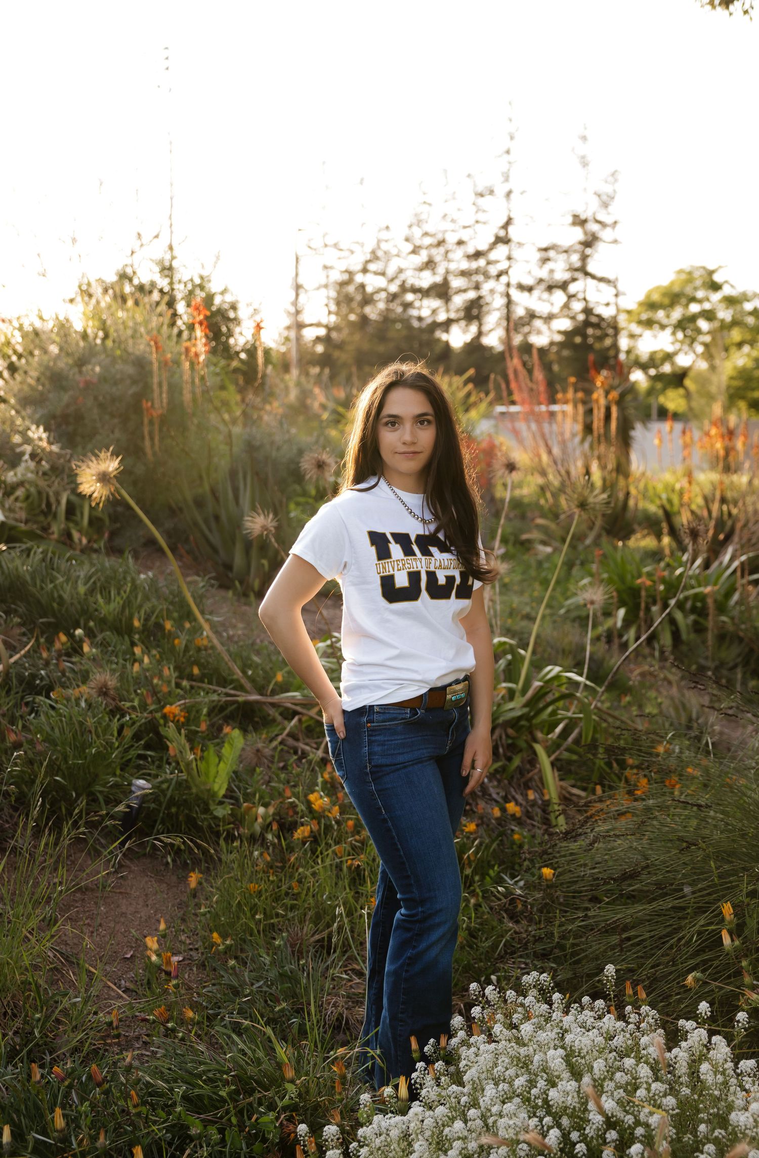 Student in white t-shirt and jeans poses in garden at golden hour.