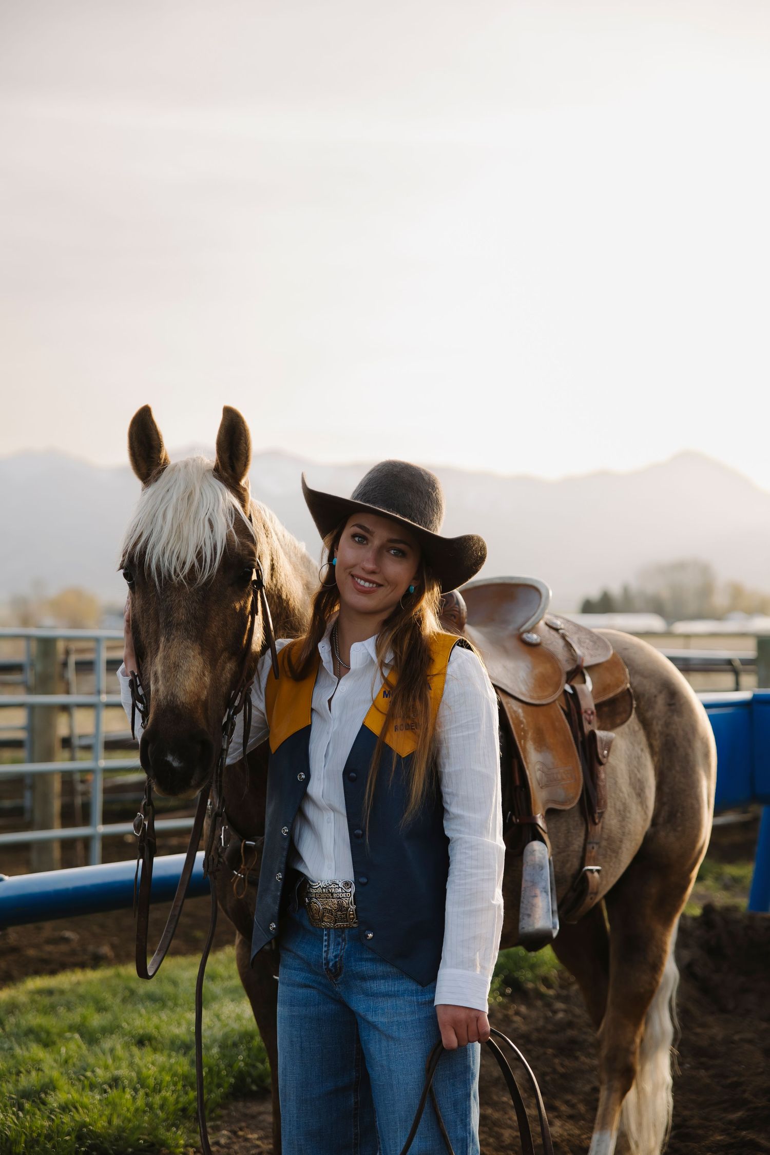 A smiling cowgirl in a hat and vest stands beside a saddled brown horse at a ranch with mountains in the background.