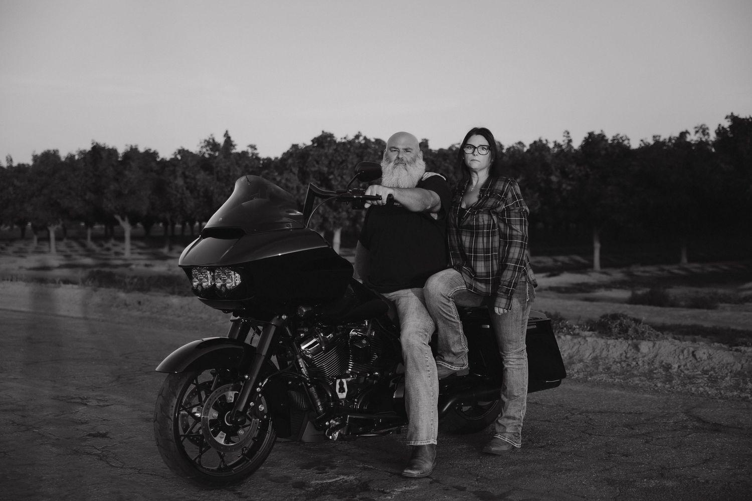 A black and white photo of a couple posing with their motorcycle in a rural setting.