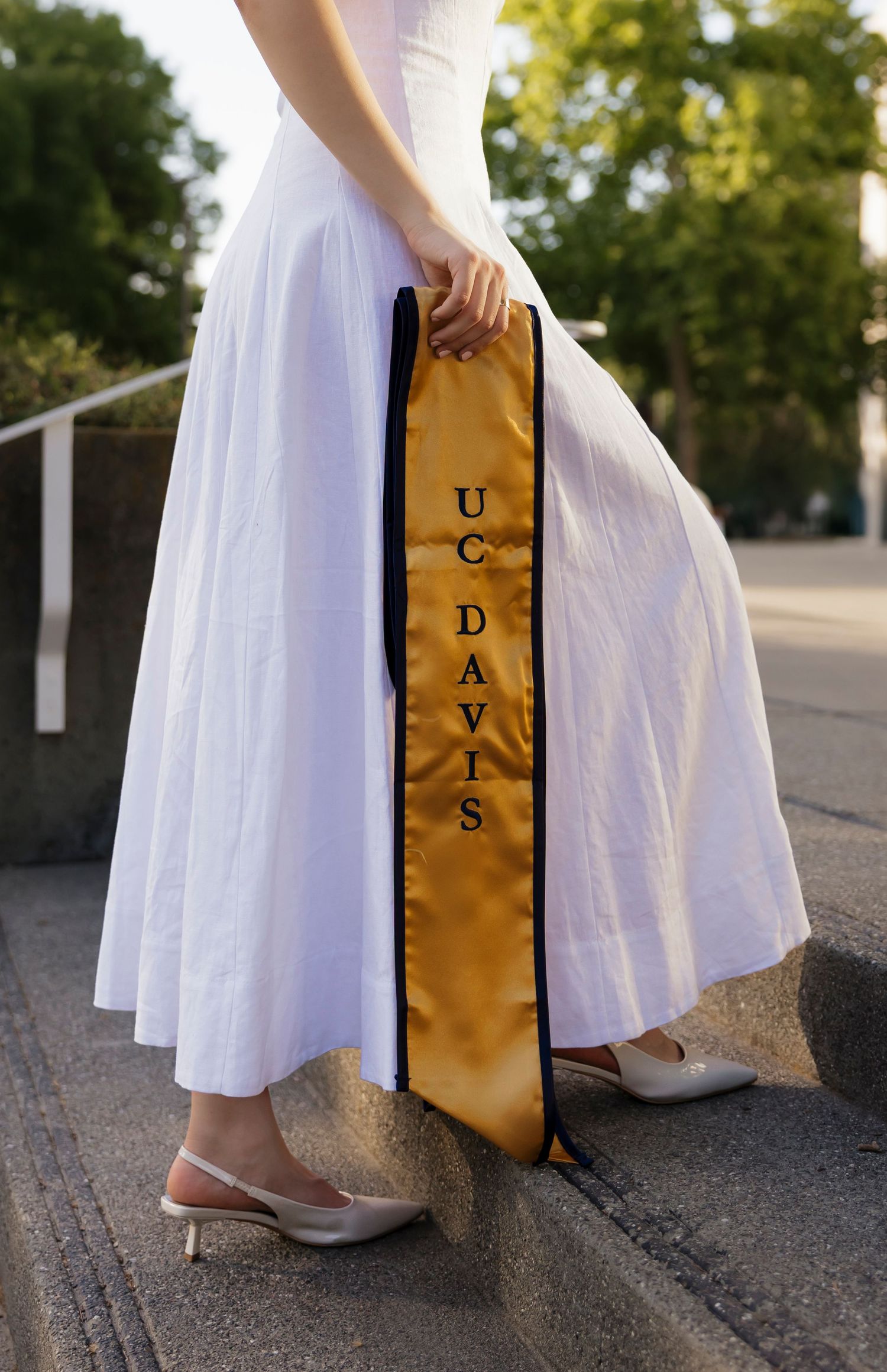 Close-up of white graduation dress and gold UC Davis sash against concrete steps.