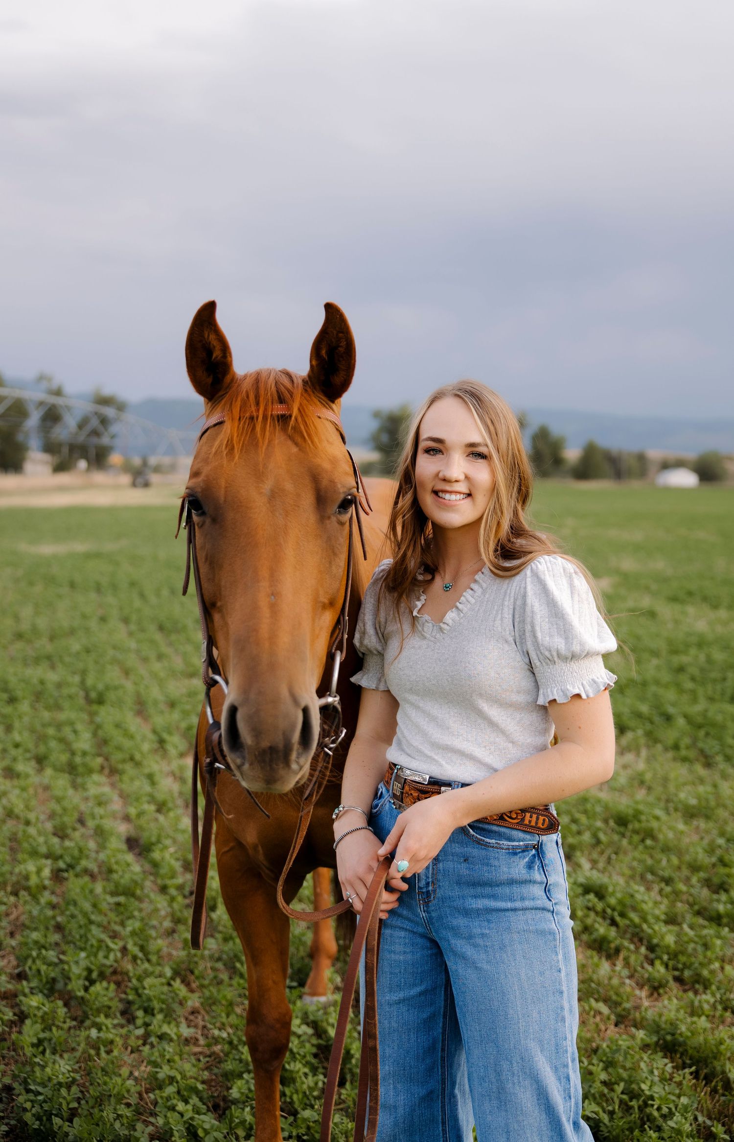 A chestnut horse stands beside someone in a green field on a cloudy day.