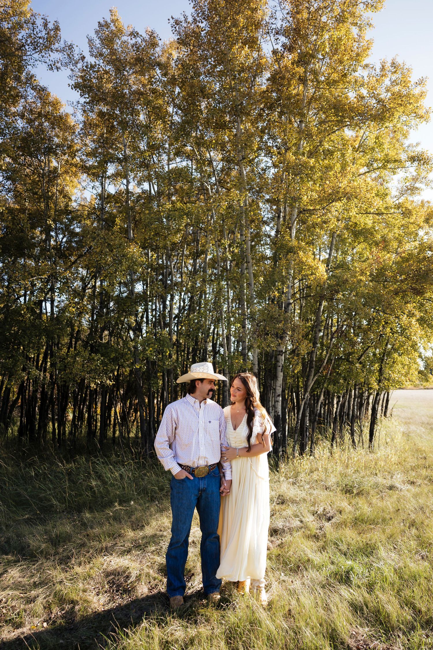 Couple stands together in front of golden aspen trees during sunset photoshoot in rustic field setting.