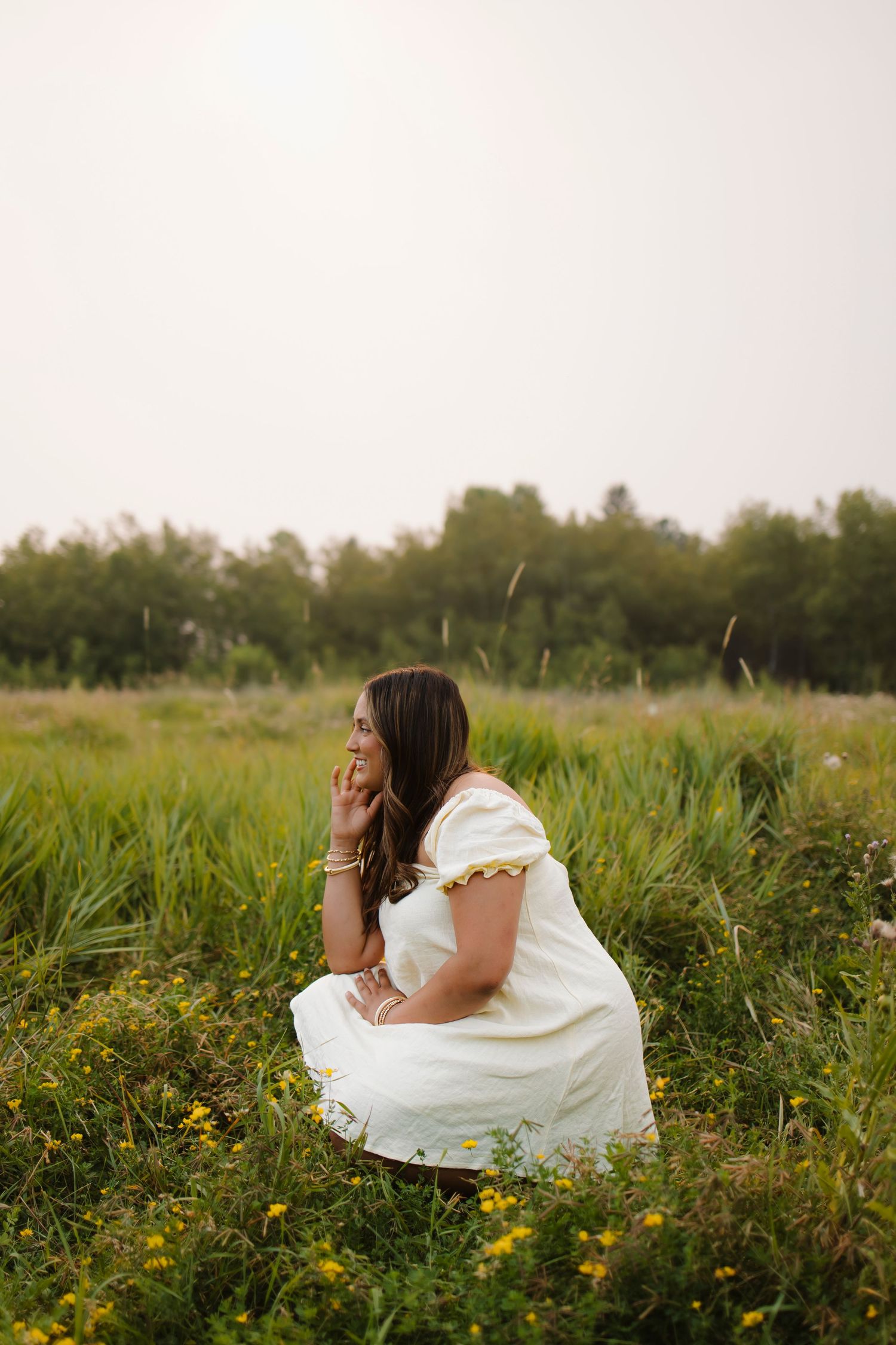 Woman in white dress crouching in a wildflower meadow, gazing thoughtfully into the distance.