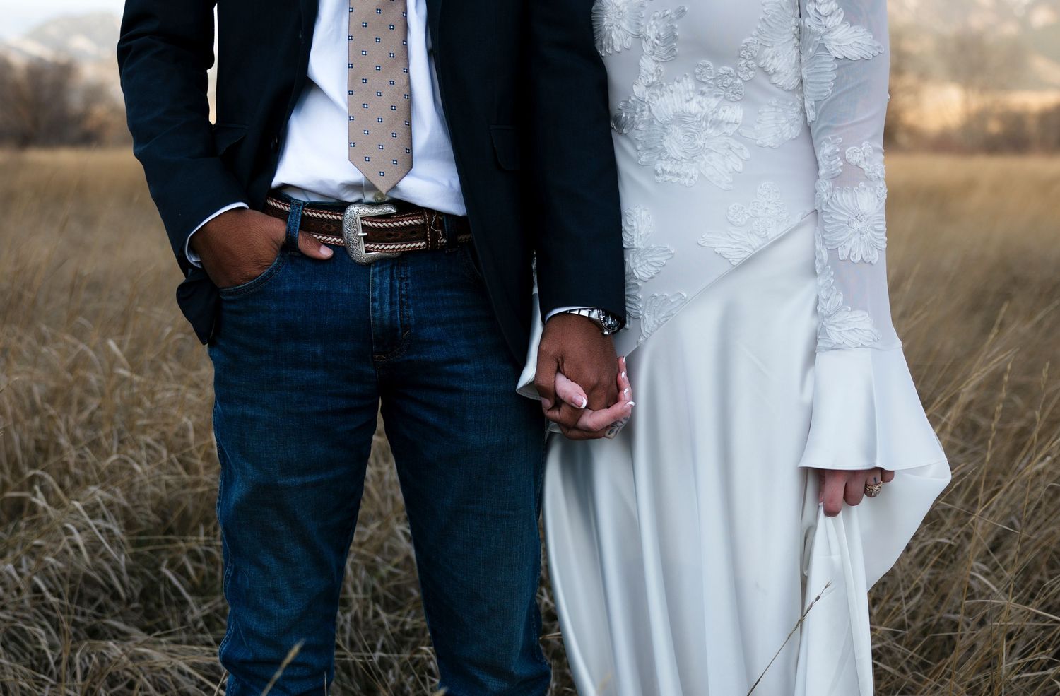 Close-up of couple holding hands in wedding attire against rustic field backdrop.