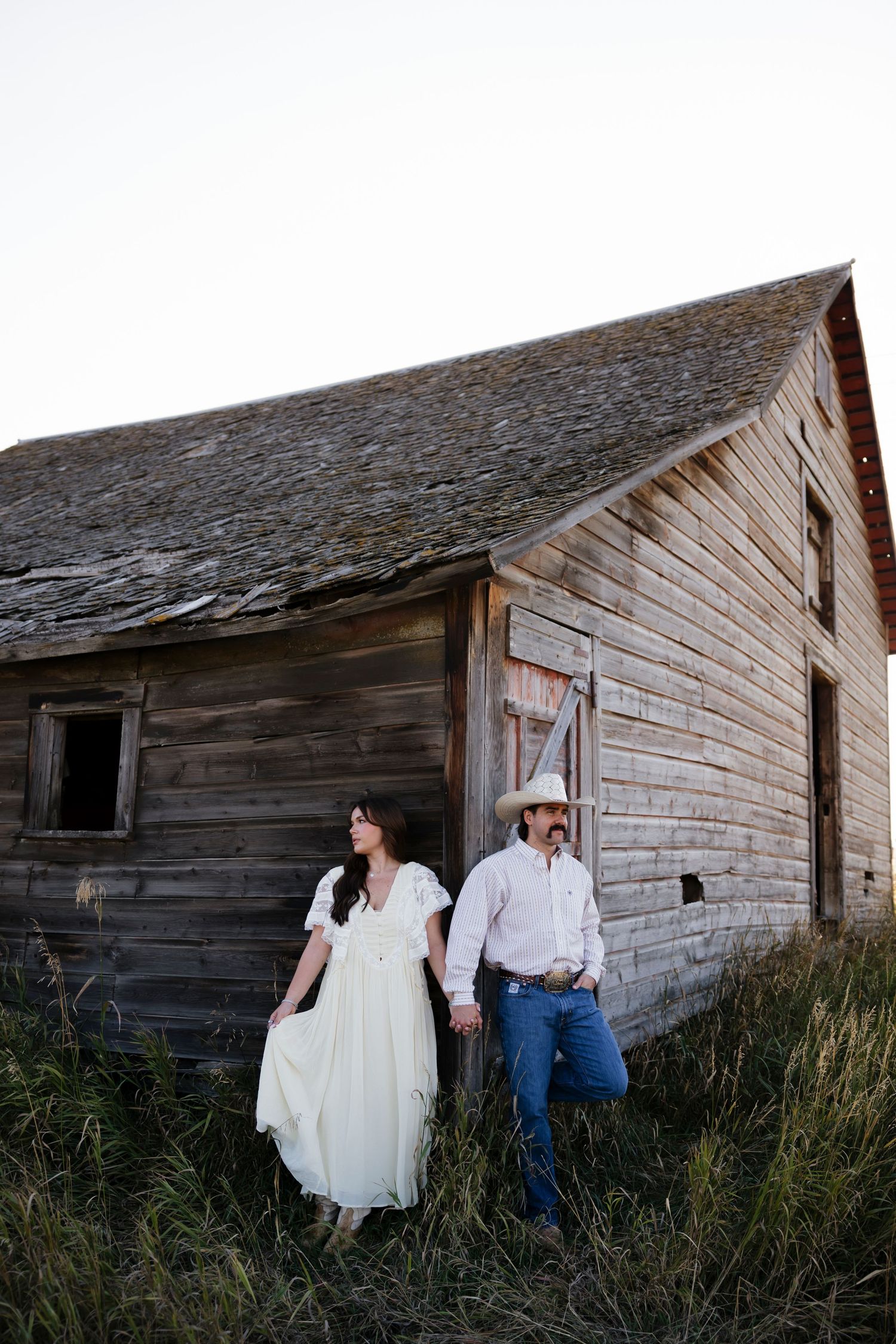 Couple poses against weathered wooden barn during rustic outdoor photoshoot.
