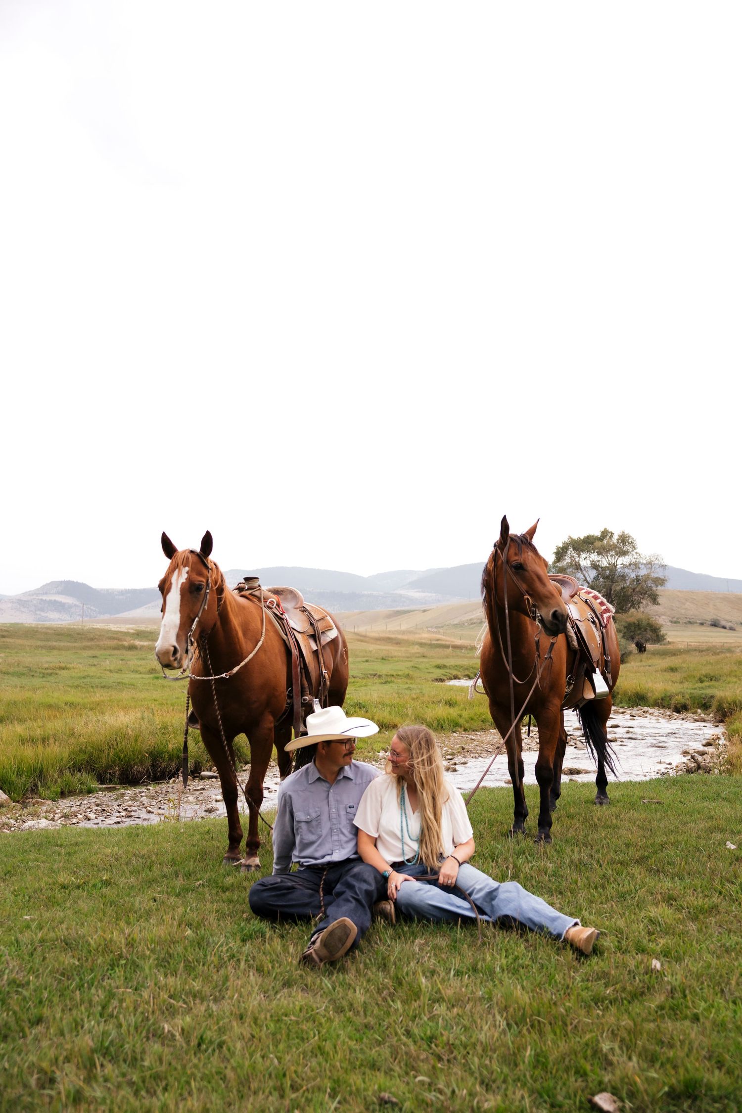 A couple relaxing in a grassy field with horses grazing nearby.