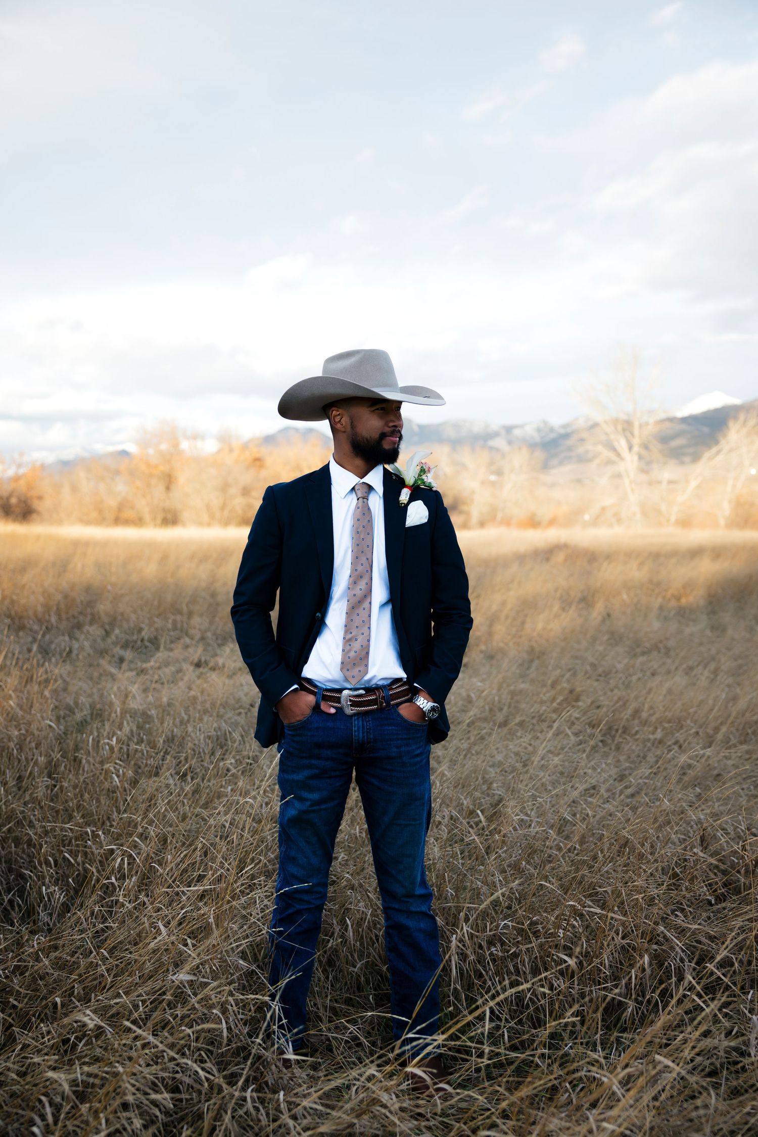 A groom in a cowboy hat and suit stands in a golden field with mountains in the background.
