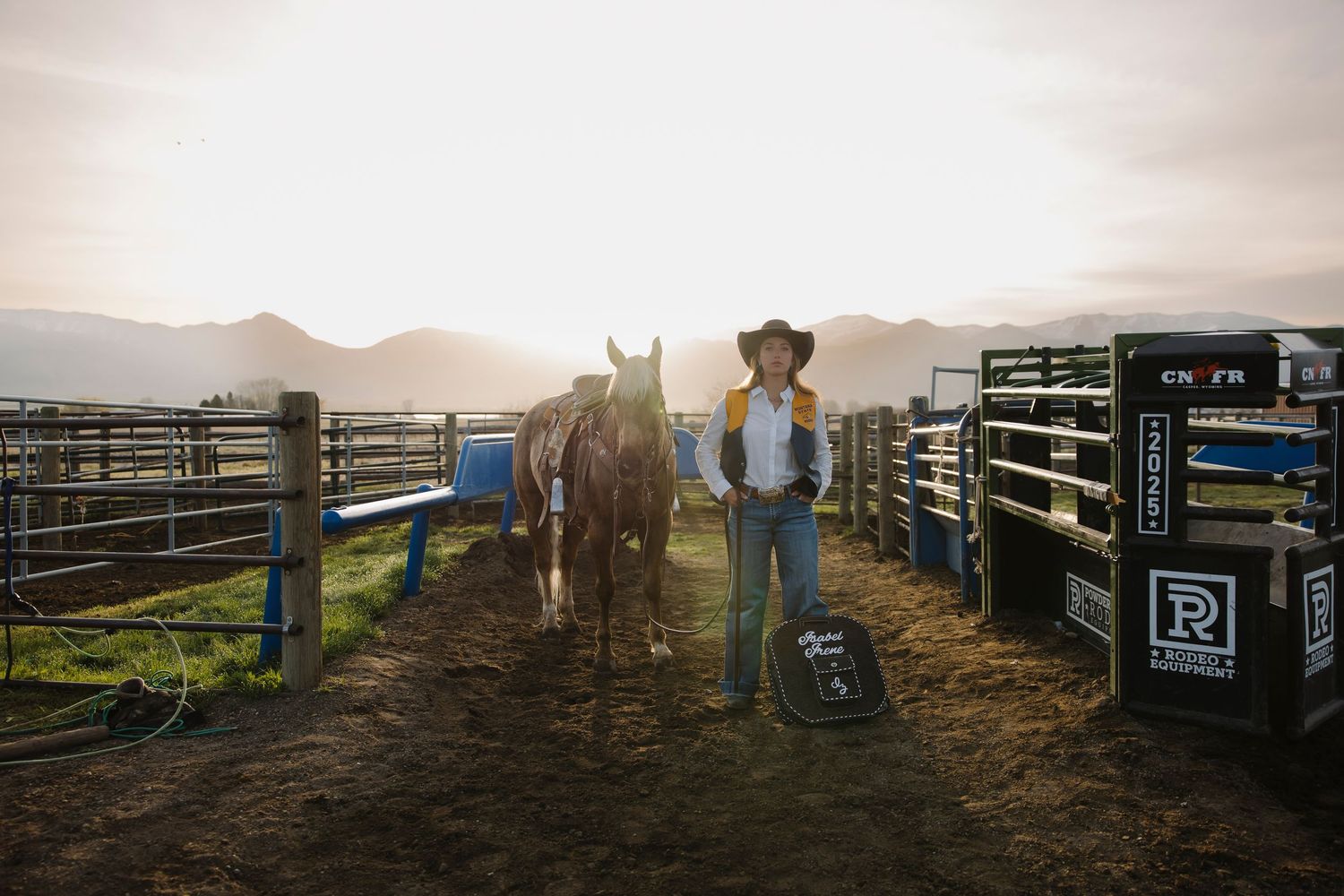 Cowboy in hat stands with brown horse at sunset near rodeo pens, mountains in background.