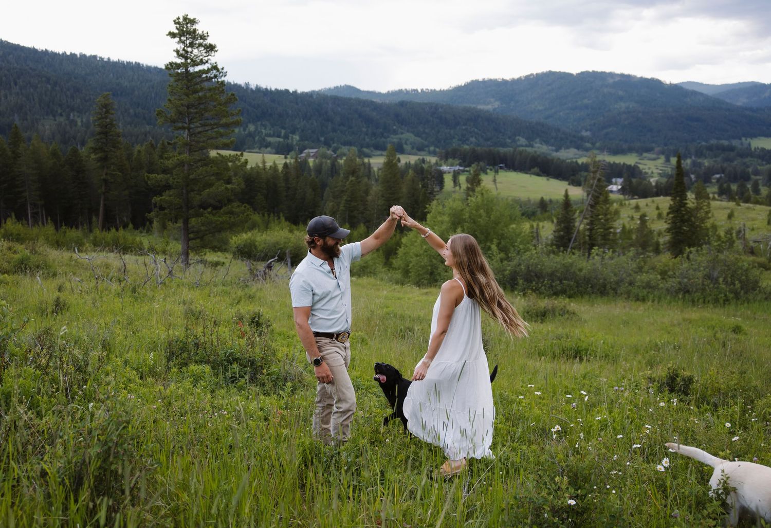 Couple dancing in a lush green meadow with mountain forest backdrop on a cloudy day.