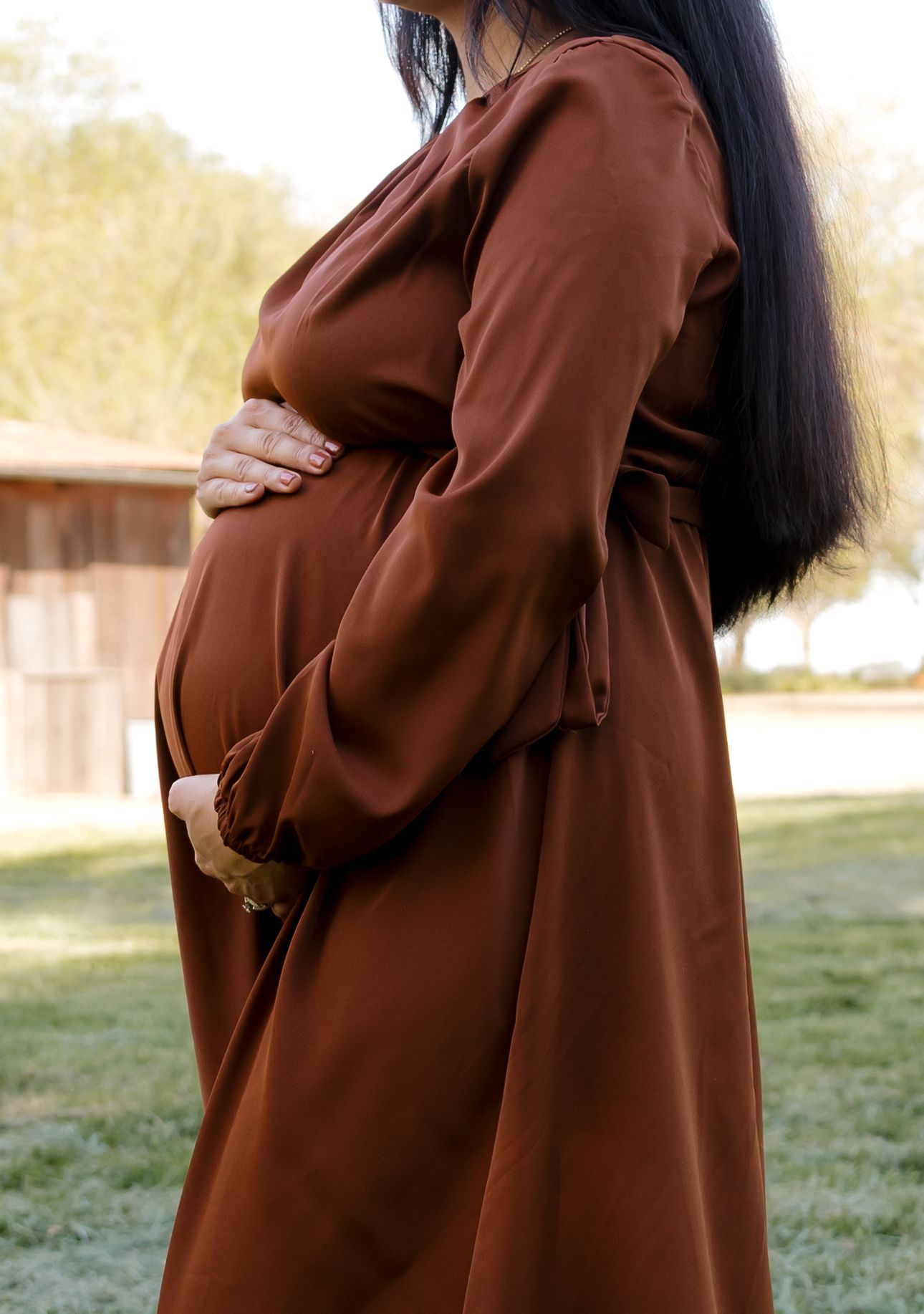 Side view of pregnant woman in brown dress cradling baby bump outdoors.