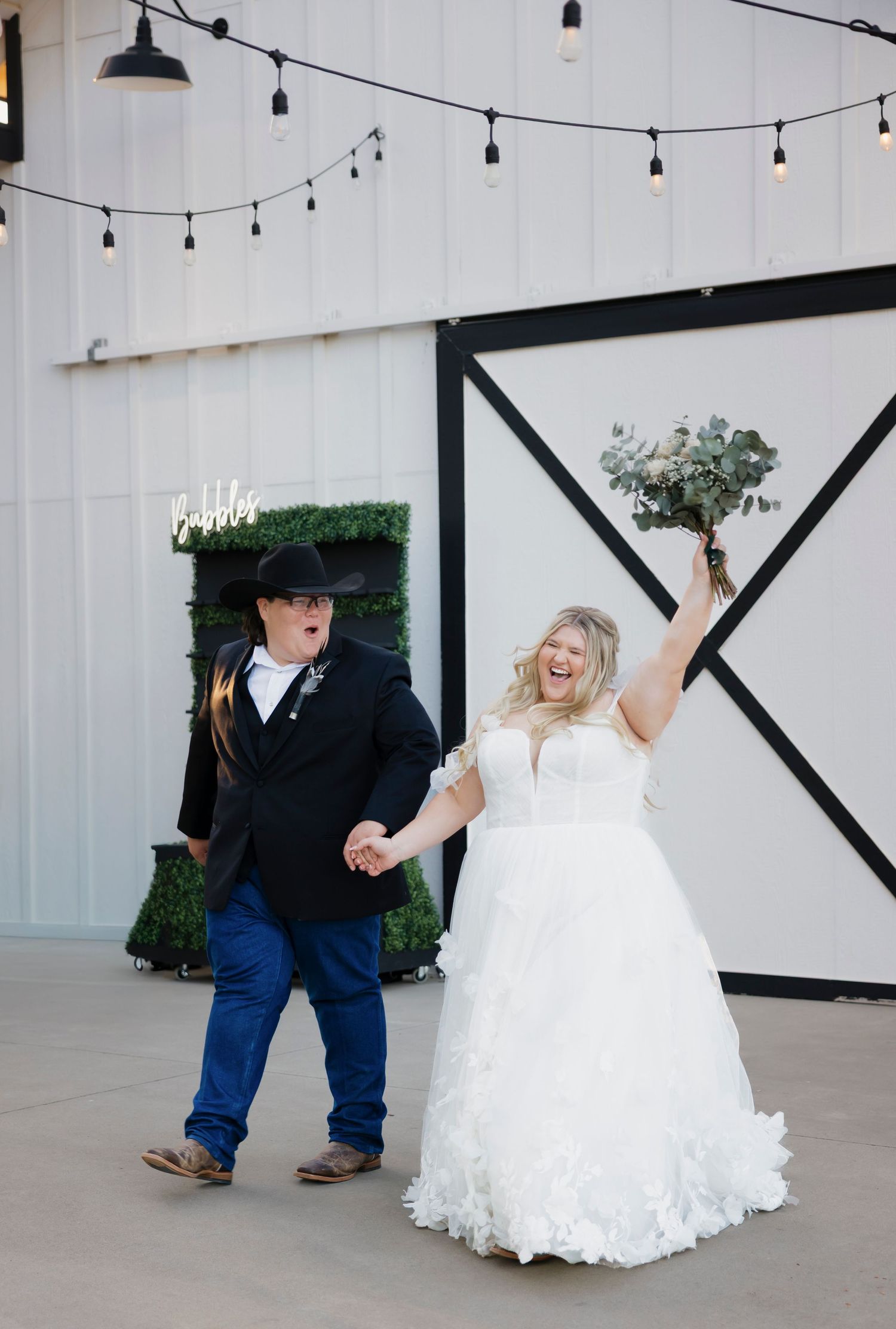 Joyful newlyweds celebrate outside a white barn venue, bride in ruffled gown holding bouquet, groom in black tux and cowboy hat.