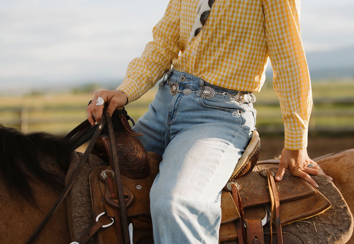 Person in yellow plaid shirt and jeans riding a brown horse with Western saddle in a rural setting.