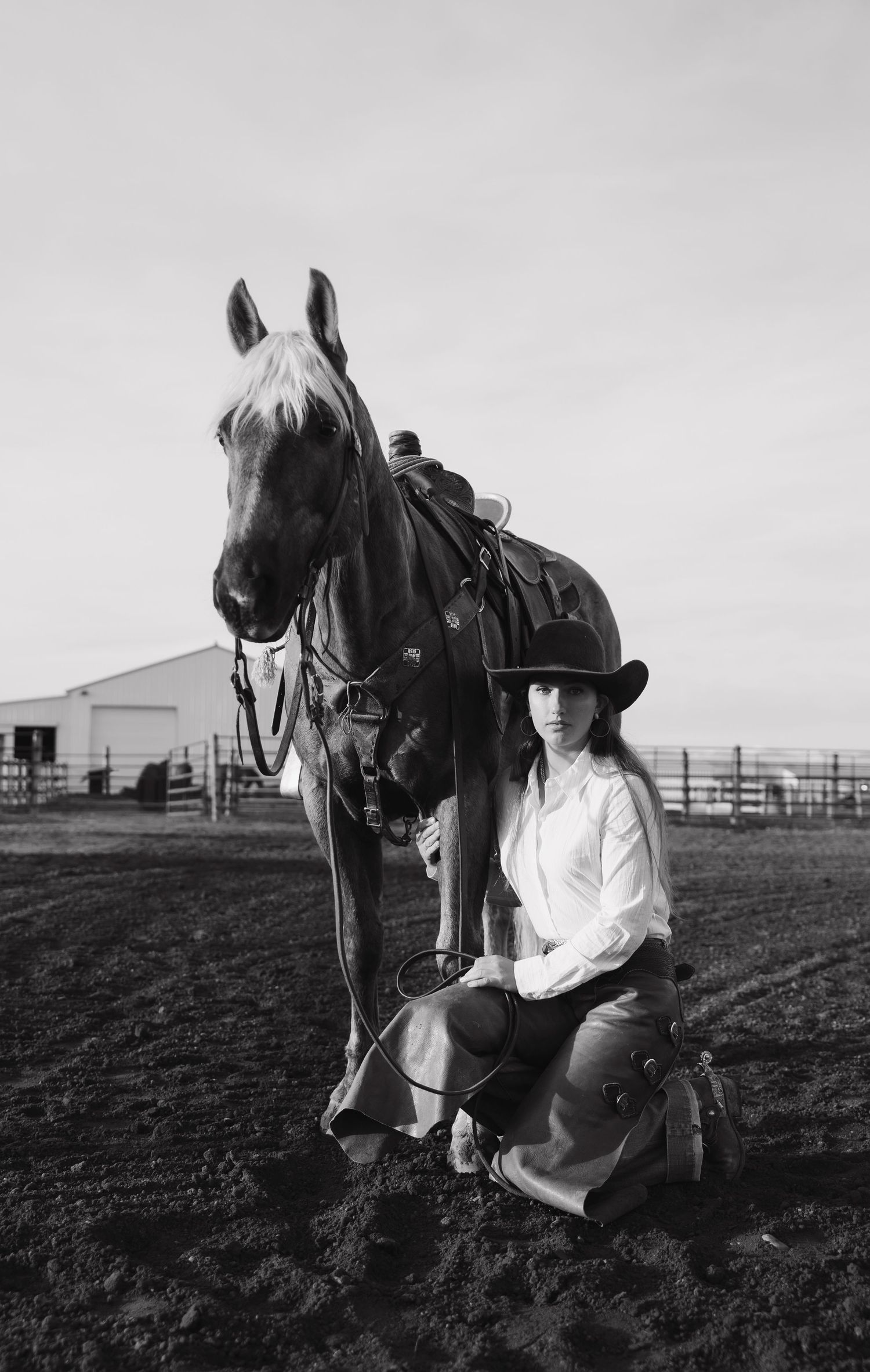 Black and white photo of a young woman in cowboy hat kneeling beside a saddled horse at a ranch.