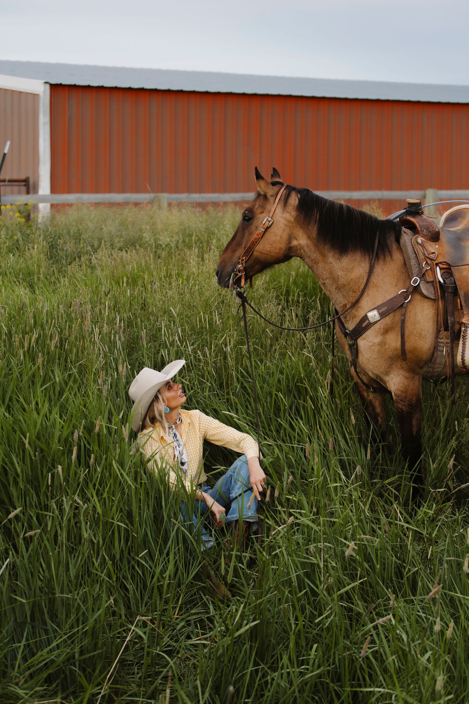Woman in cowboy hat sits in tall grass beside a saddled buckskin horse near a red barn.