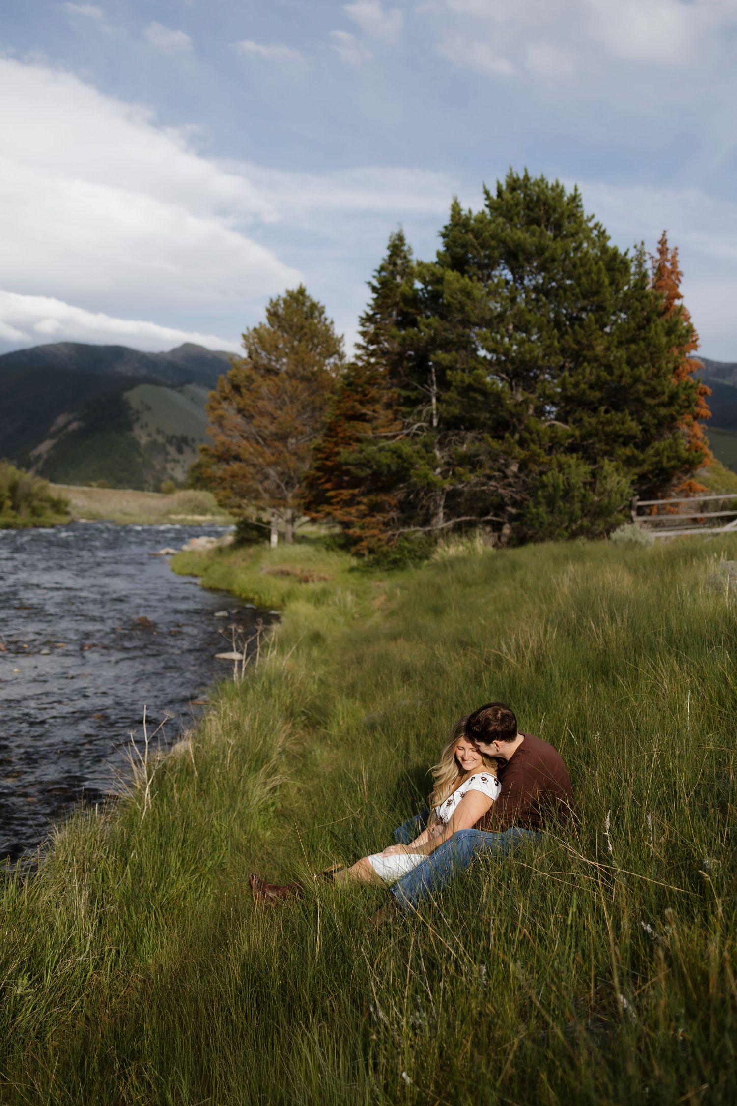Couple sitting romantically in tall grass beside a mountain river, surrounded by pine trees and rolling hills.