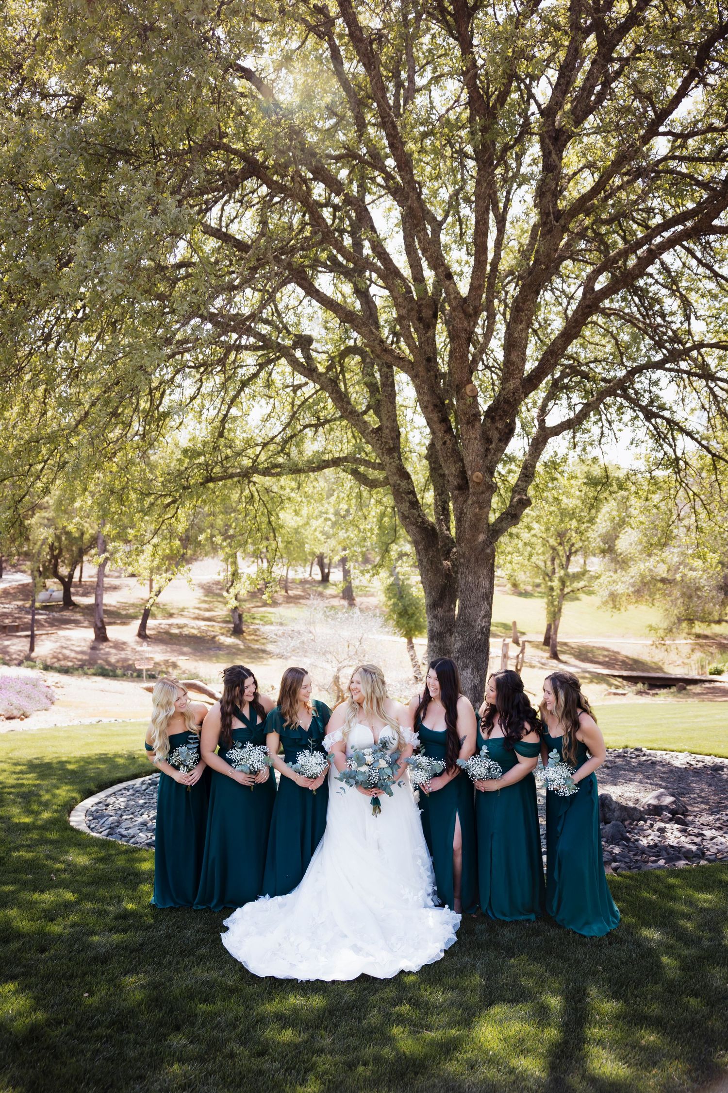 Bride in white gown with bridesmaids in emerald green dresses holding bouquets under a large oak tree outdoors.