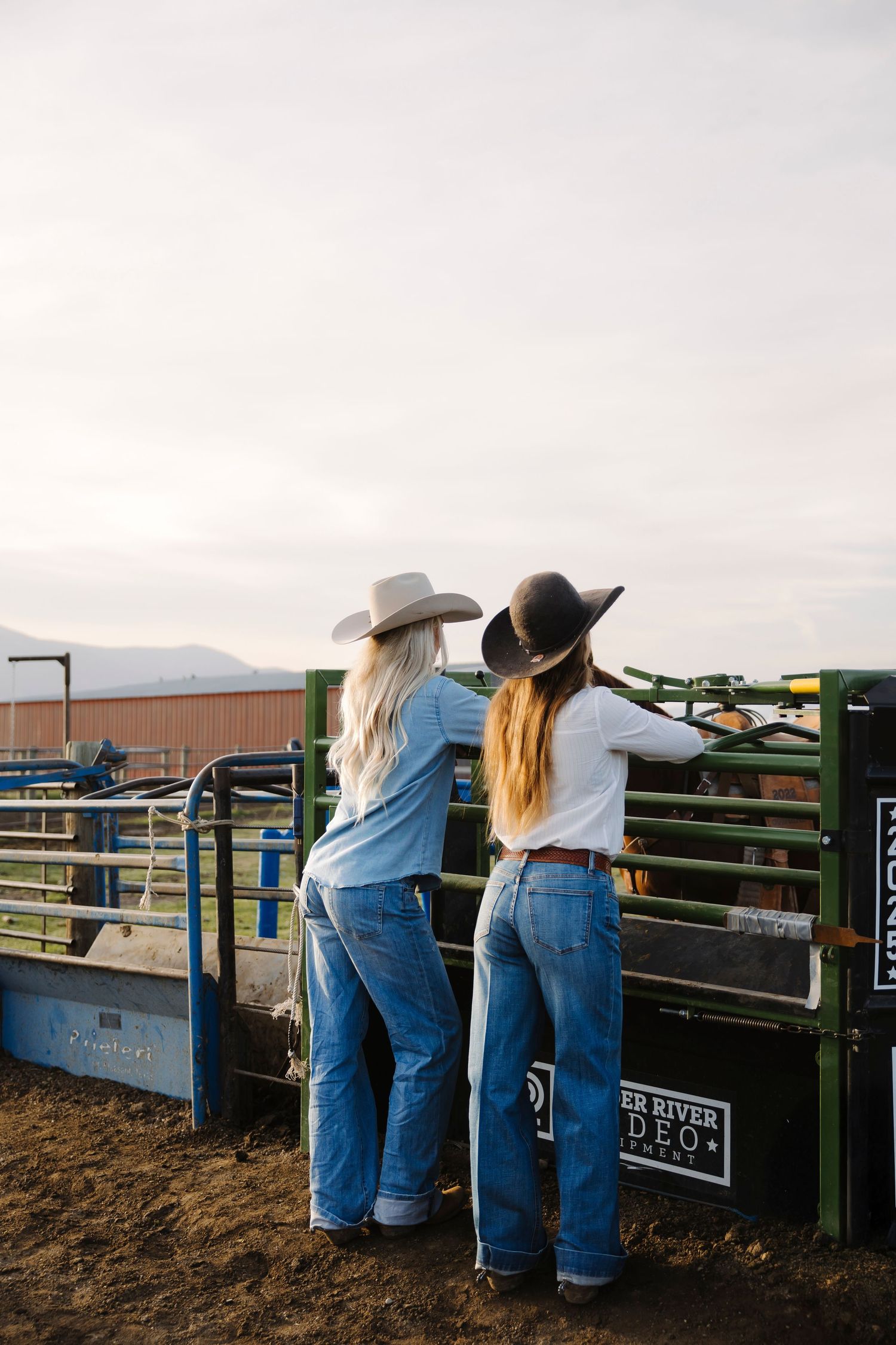 Two women in cowboy hats and jeans leaning on rodeo gates at an outdoor arena during golden hour.