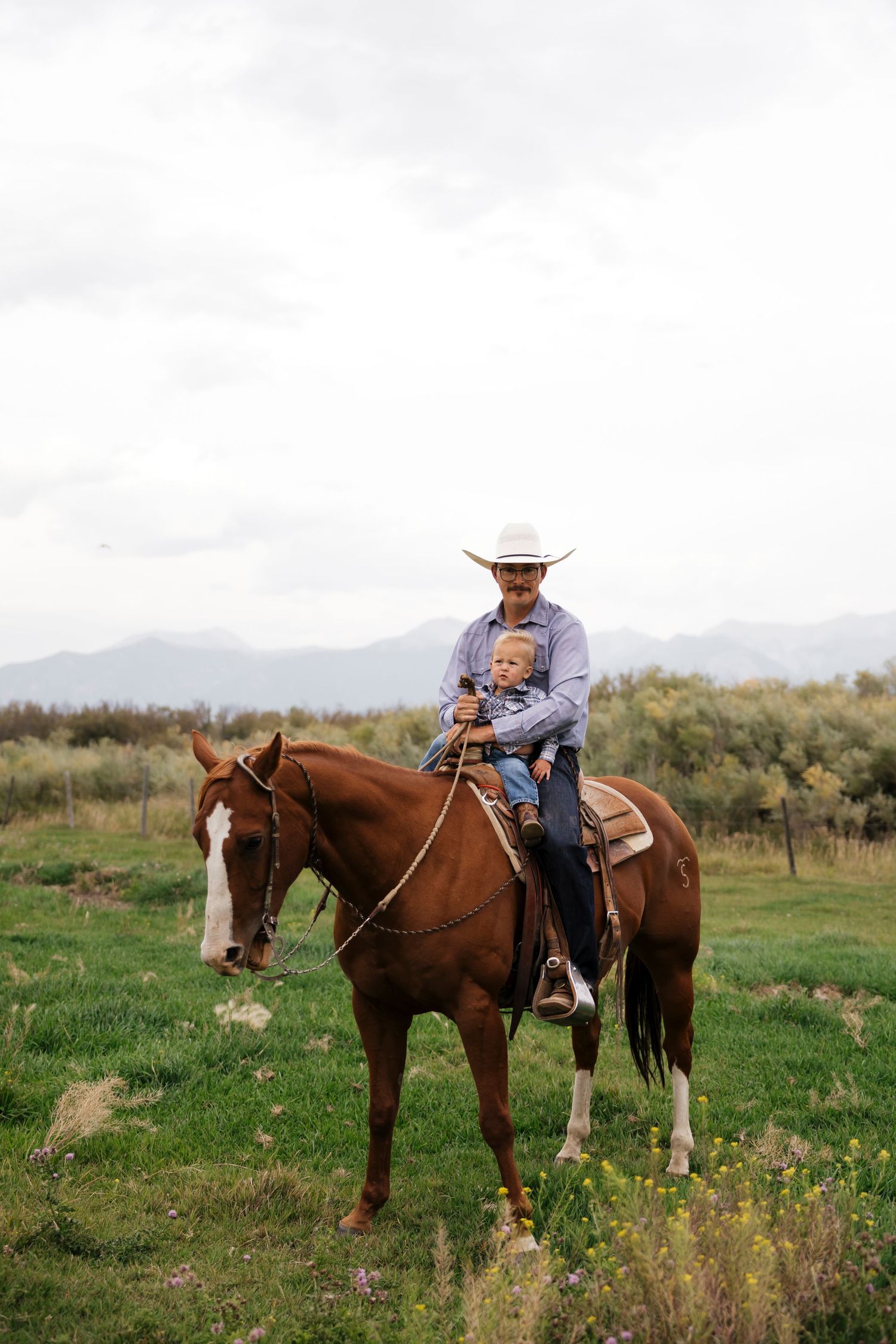 A horseback rider in western attire guides their horse through a grassy meadow landscape.