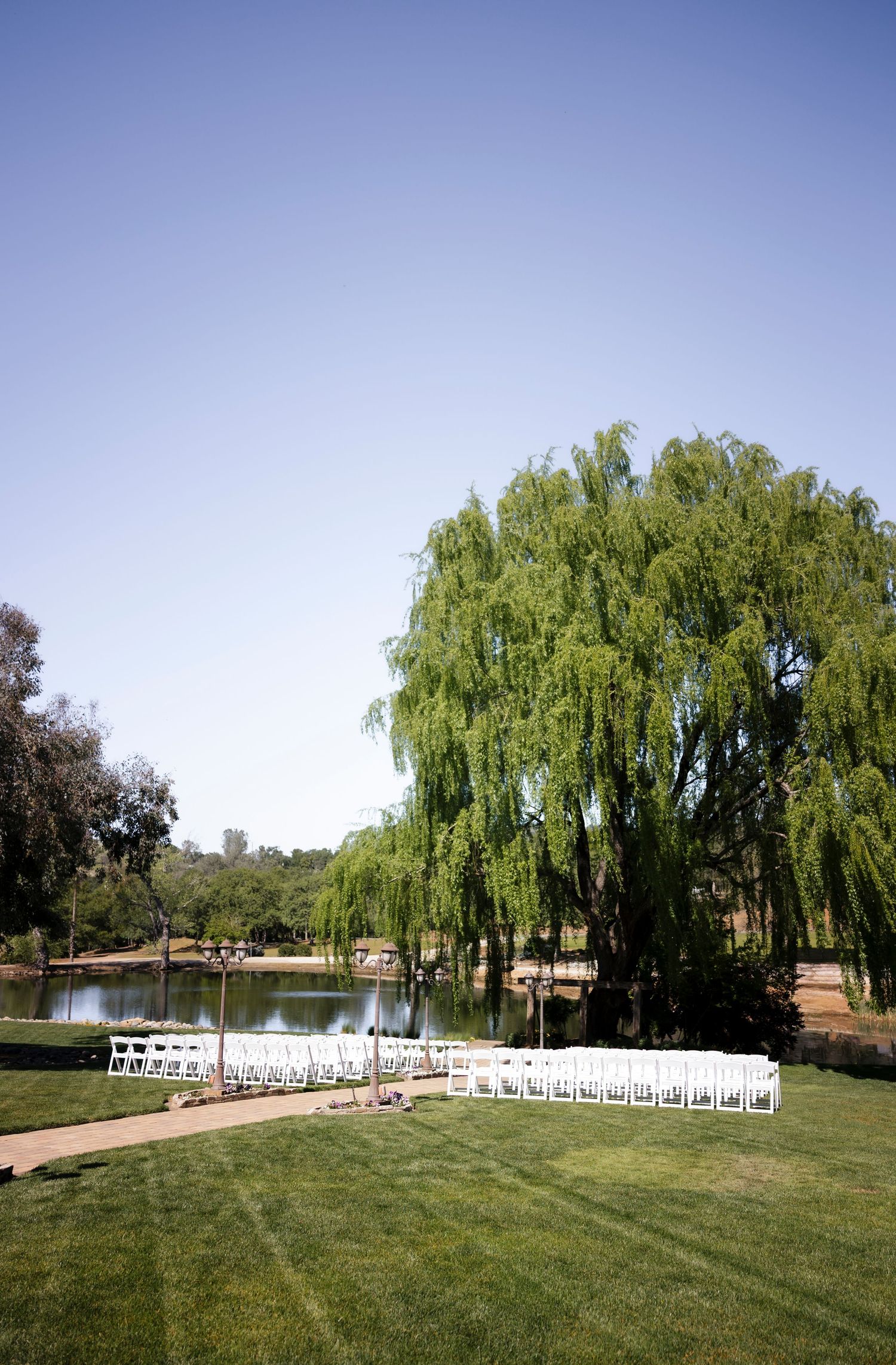 Outdoor wedding ceremony setup with white chairs facing a lake under drooping willow trees.
