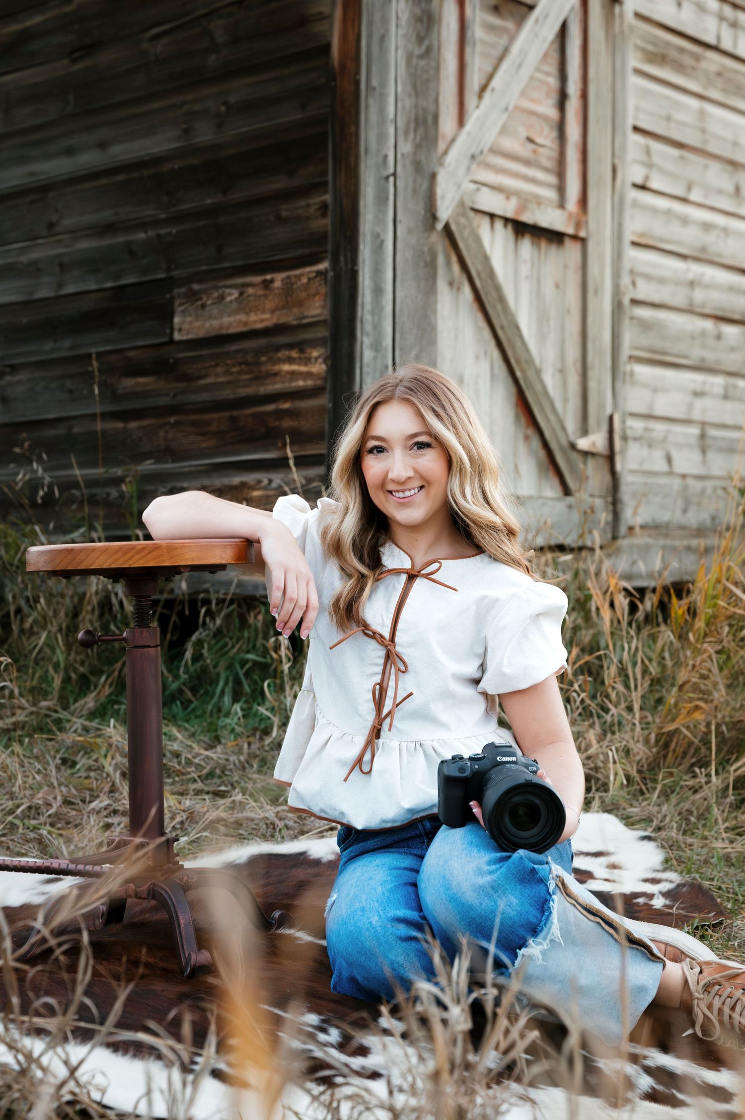 Photographer with camera sits casually by weathered barn wall surrounded by tall grass.