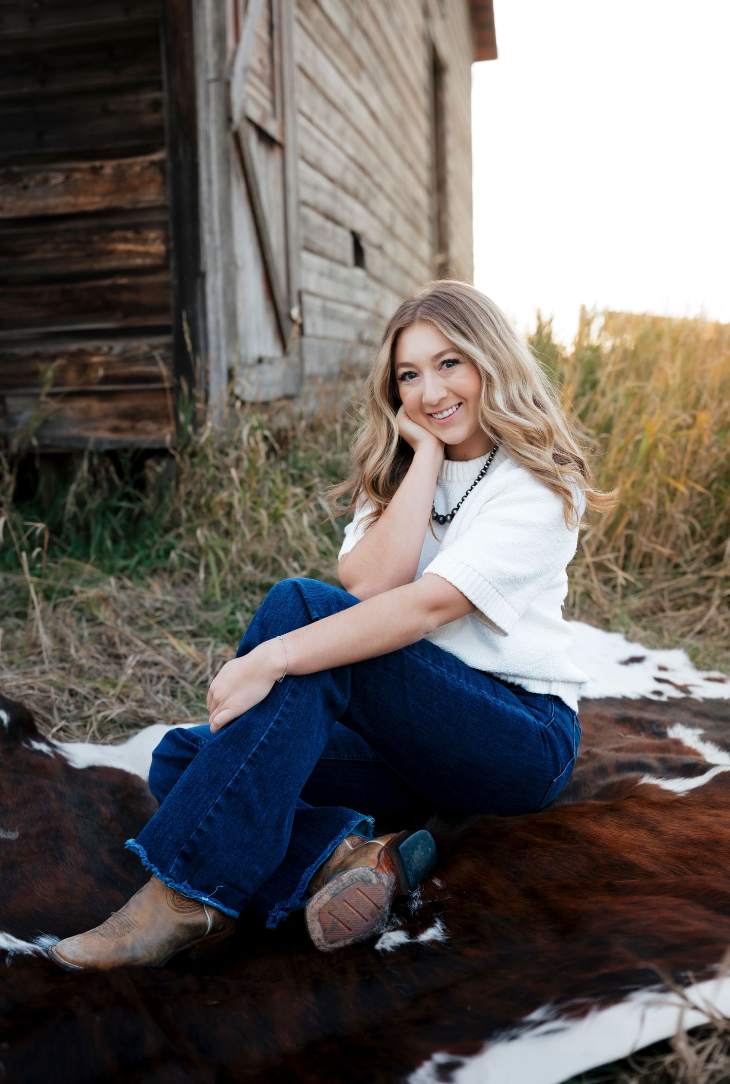 Photographer relaxes on cowhide blanket against rustic barn backdrop in casual country attire.