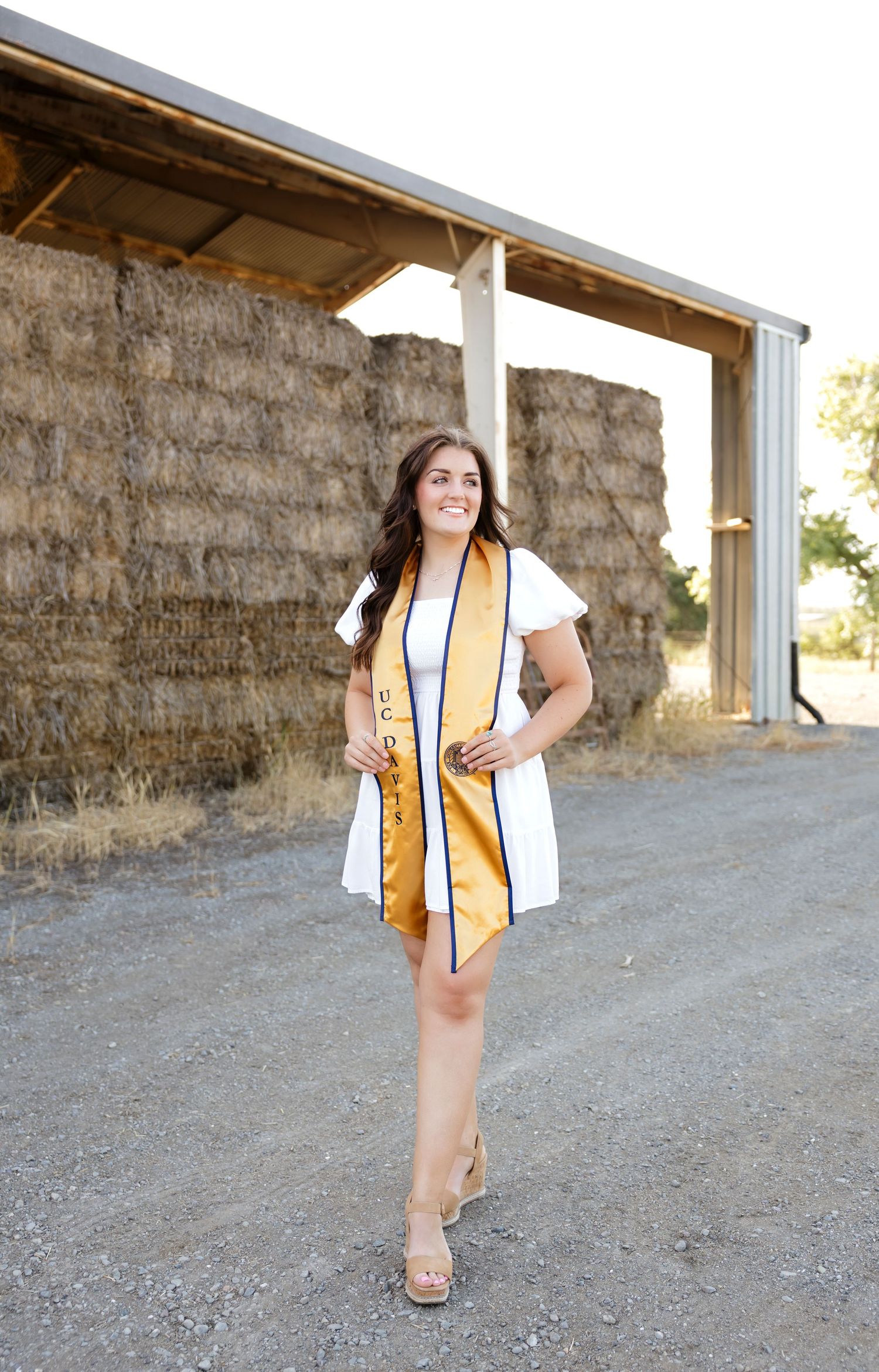 Graduate in white dress and gold sash poses in front of rustic barn wall.