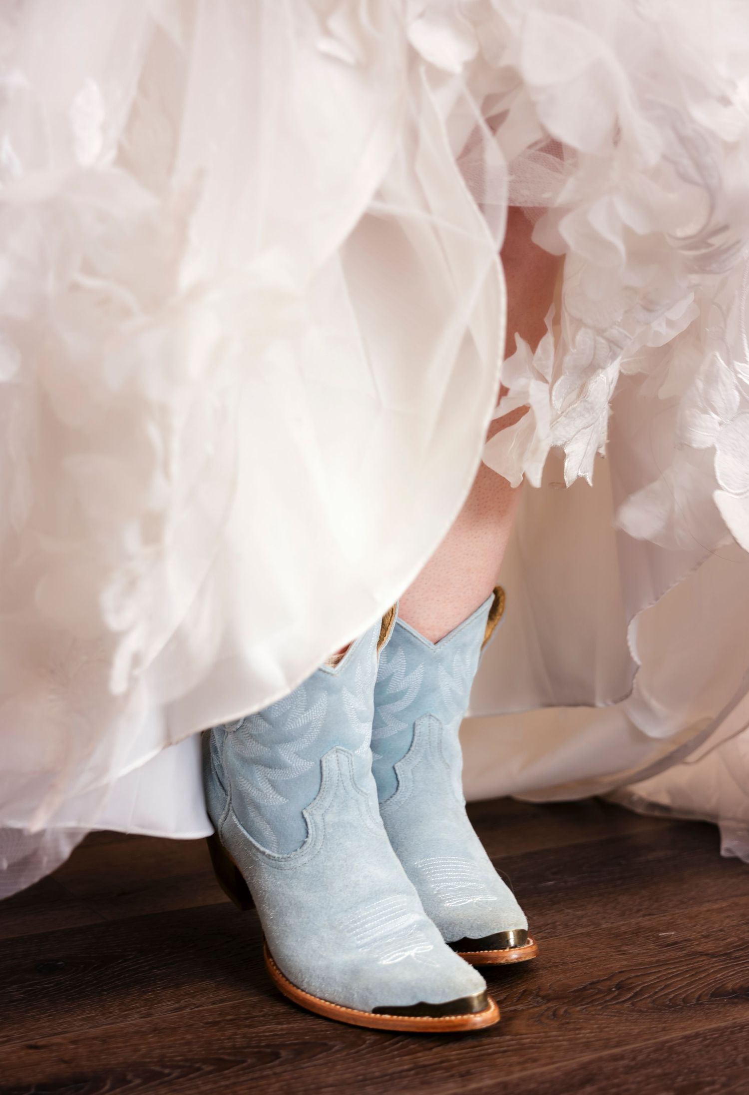 Light blue cowboy boots peek out beneath layers of white wedding dress fabric on a wooden floor.