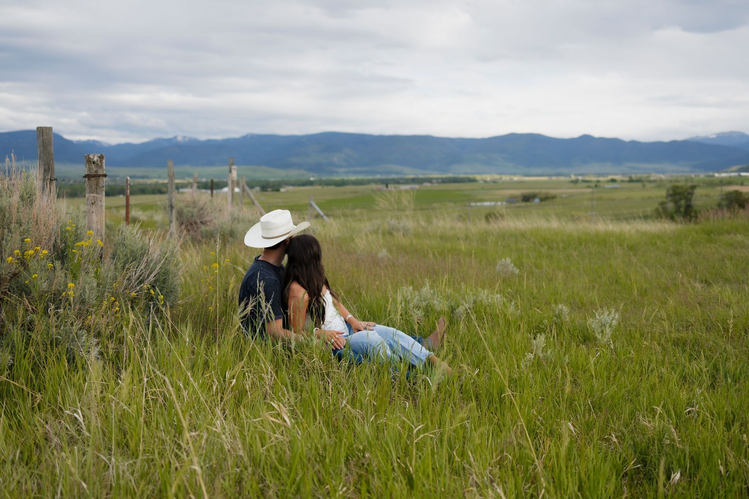 Couple kissing in tall green grass with mountain backdrop under cloudy skies in a scenic rural landscape.
