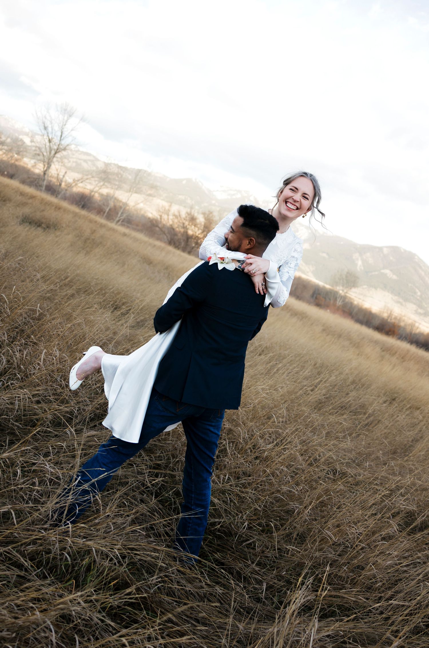 A joyful groom lifts his smiling bride in a white dress in a golden field with mountains in the background.