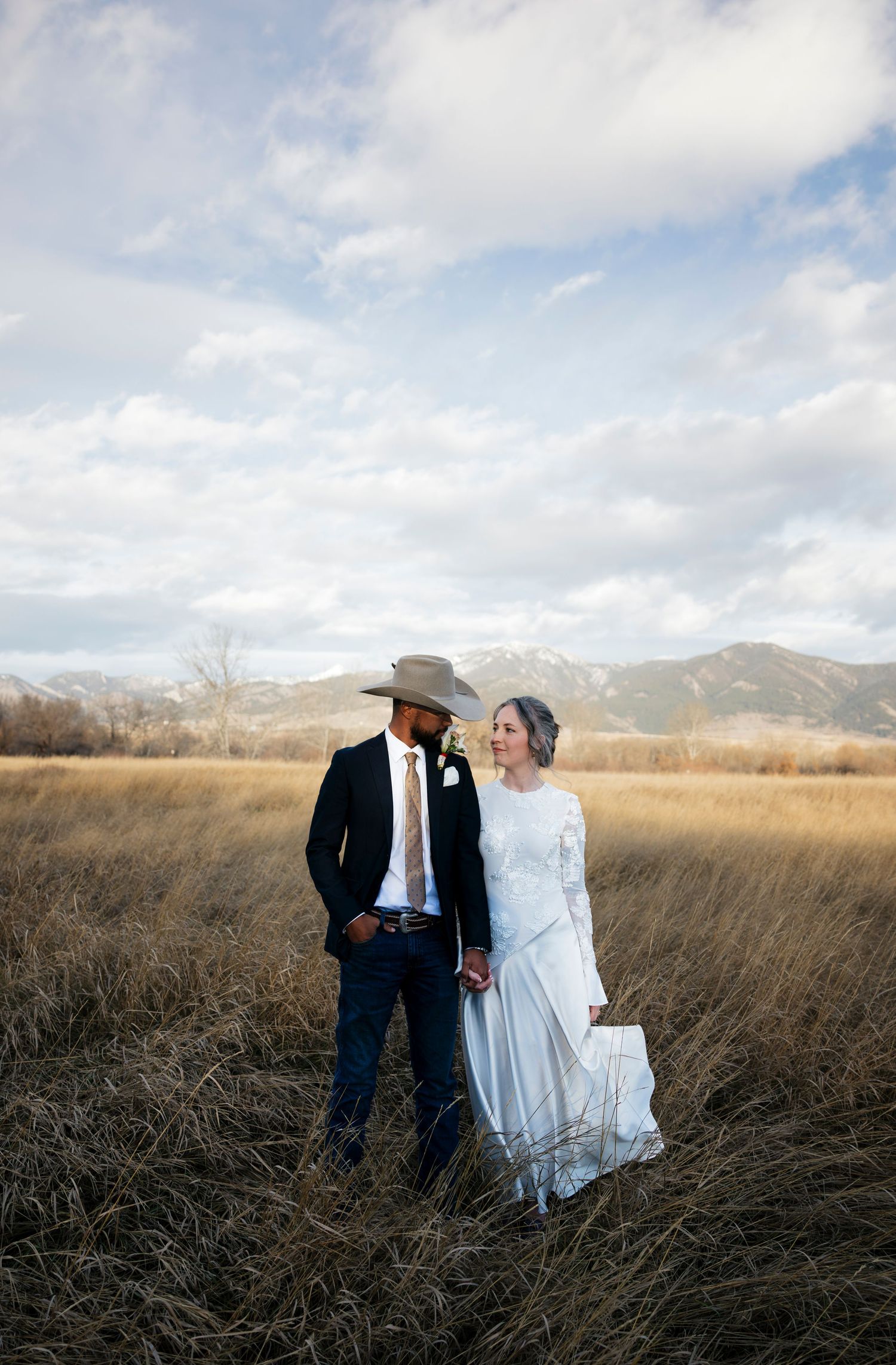 Couple in western wedding attire standing in tall grass field with mountains in background.