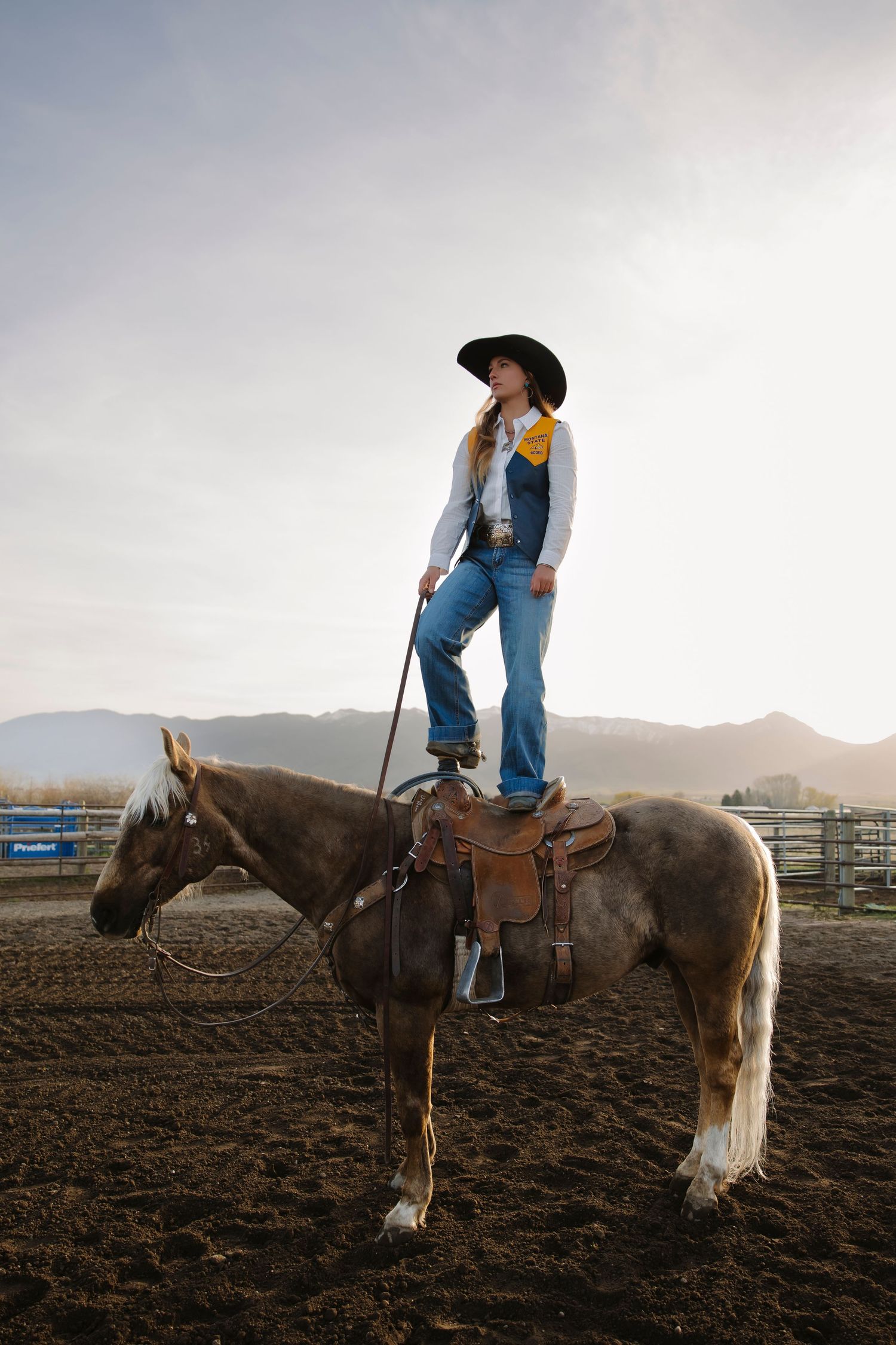 Cowgirl in black hat and jeans stands atop a saddled horse at a rodeo arena with mountains in background.