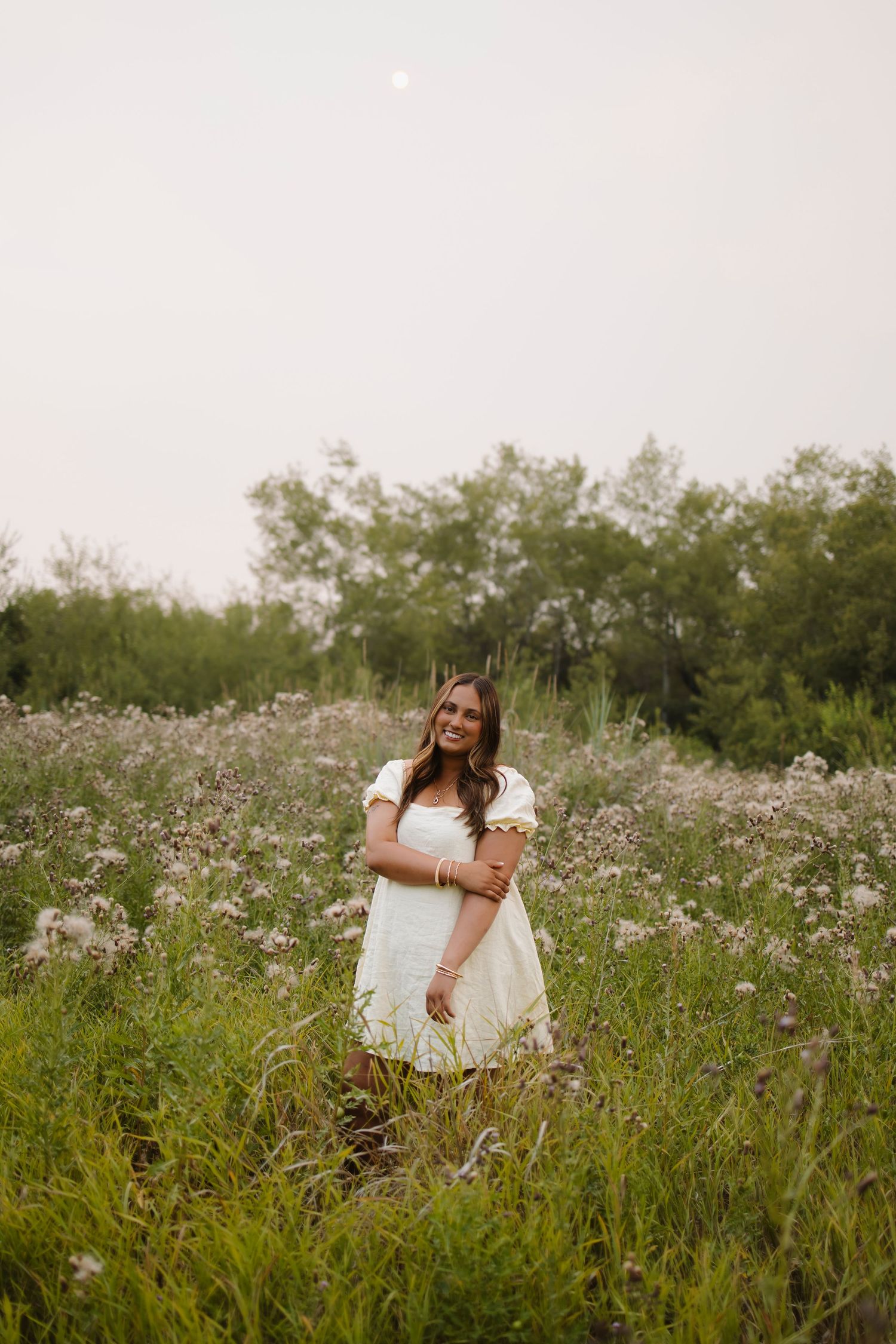 A smiling woman in a white dress stands in a lush wildflower field surrounded by green trees.