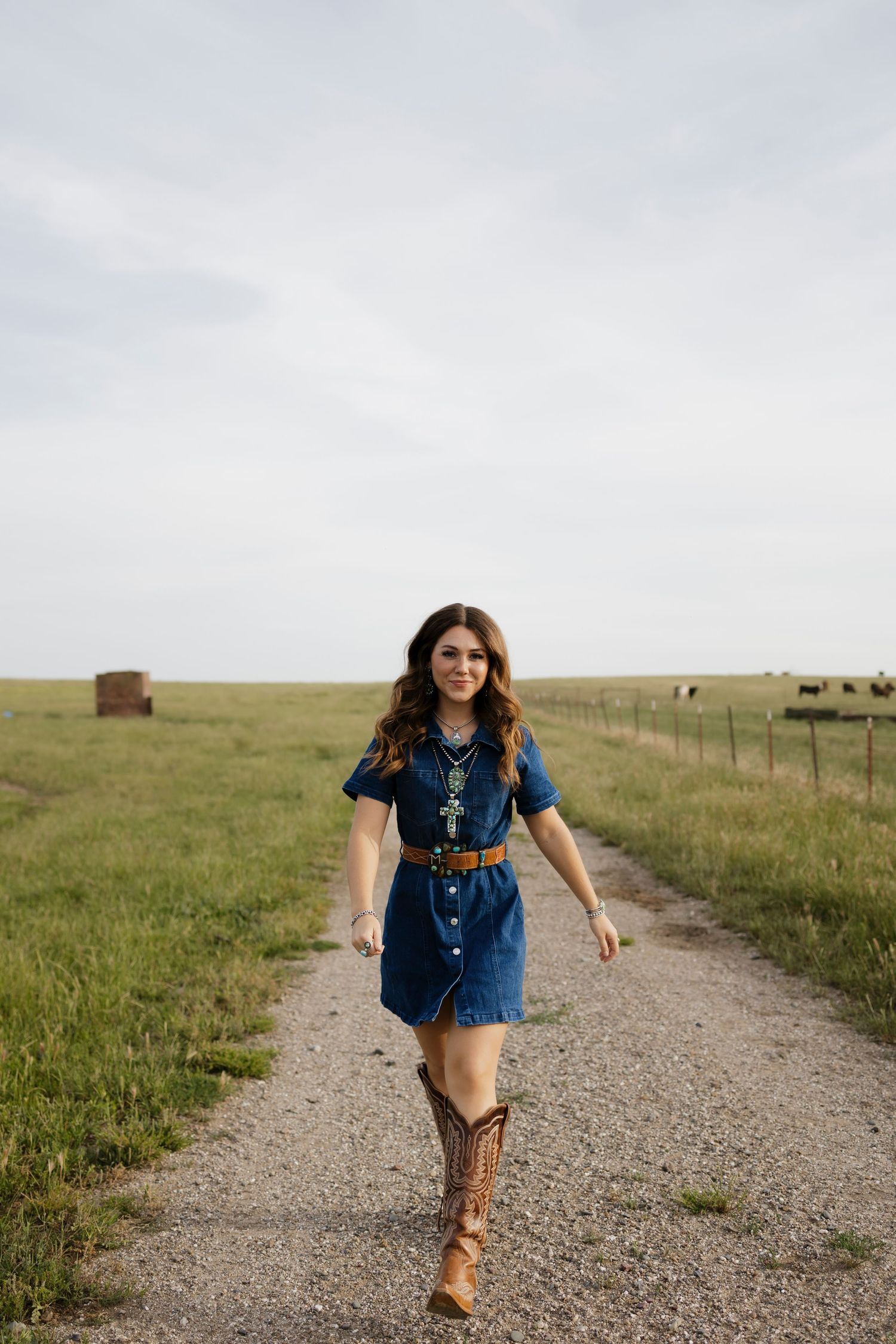 Young woman in denim dress, cowboy boots, and cross necklace walking down a rural gravel path in a green field.