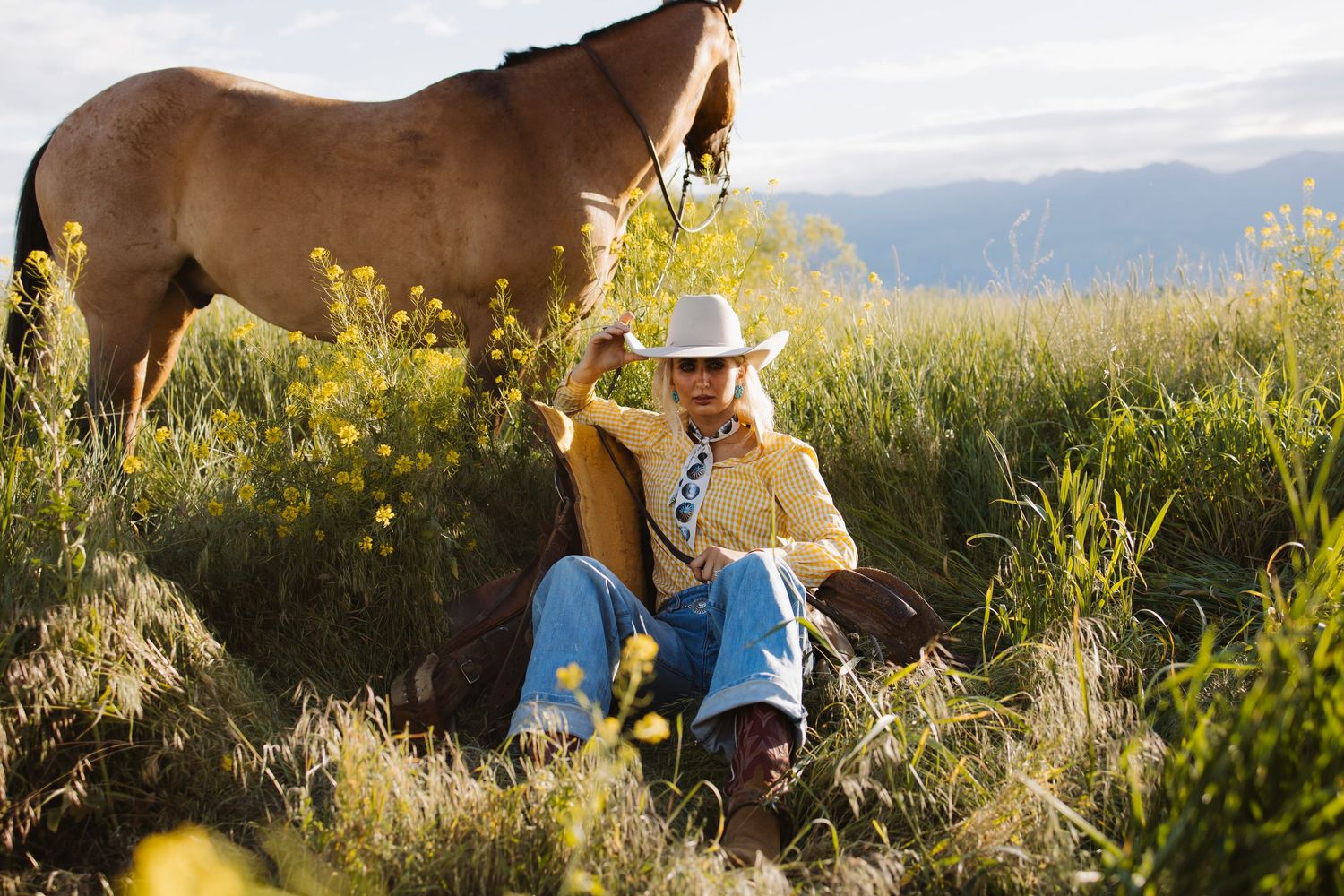 Cowboy in yellow jacket and white hat sits in wildflower field beside a brown horse with mountain views.