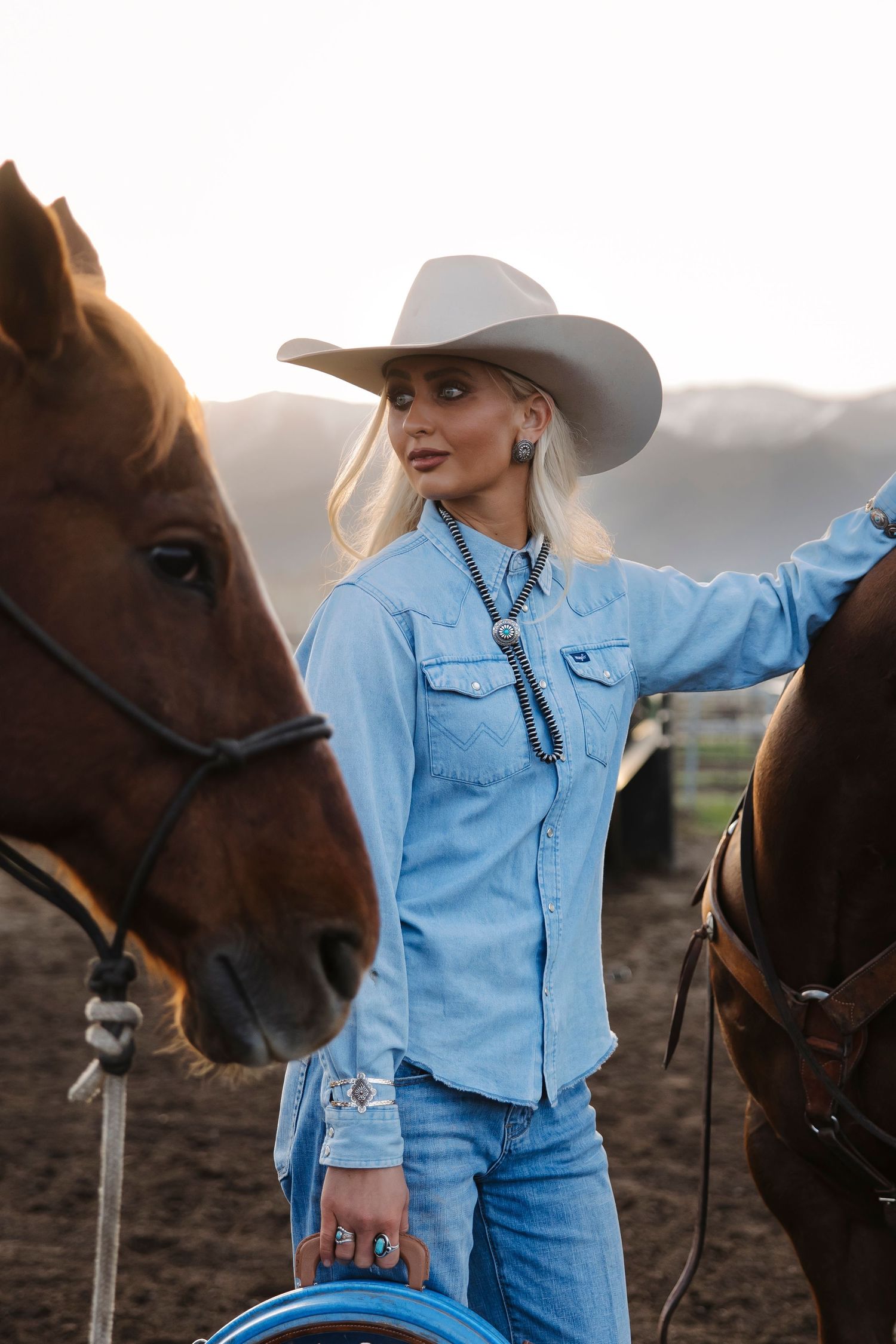 Blonde woman in cowboy hat and denim outfit stands between two brown horses at a ranch during golden hour.