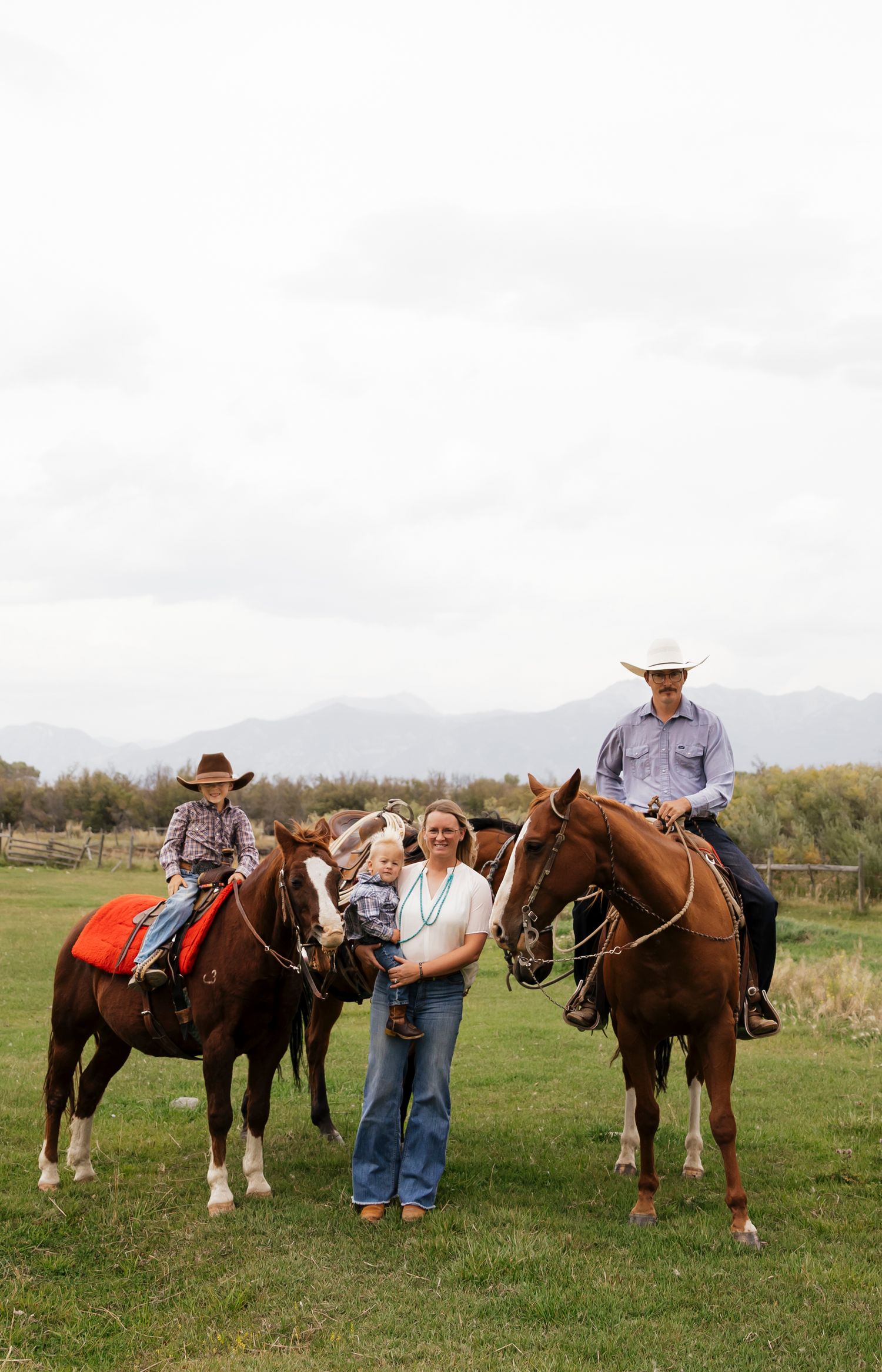 Line of horses and riders in western attire standing in a grassy field with mountains visible.