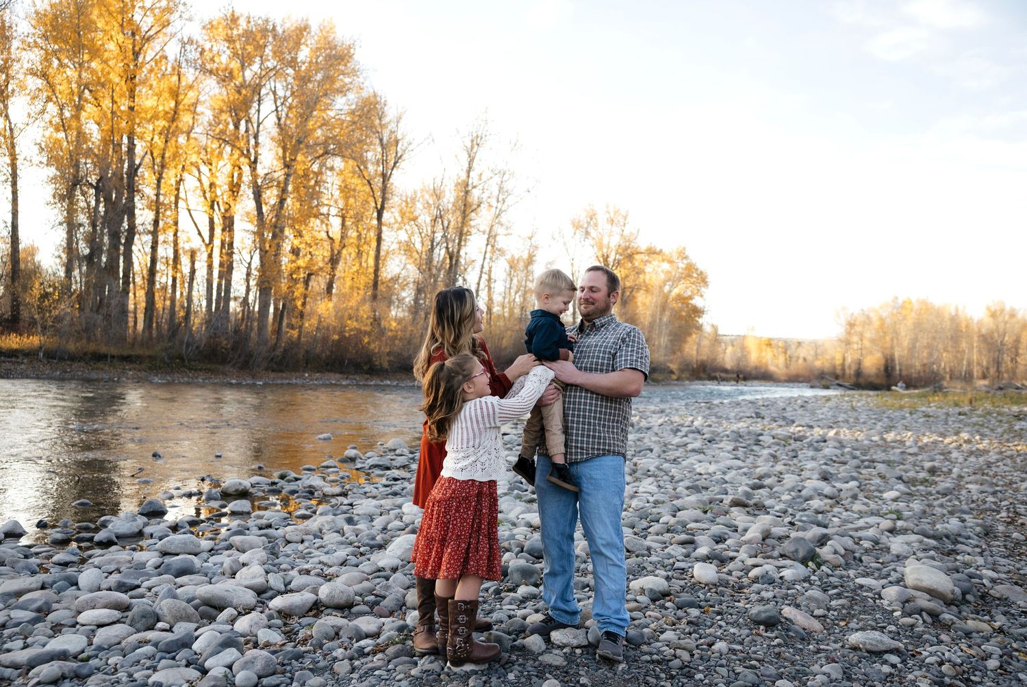 Family enjoying autumn scenery on a rocky riverbank with golden trees in the background.