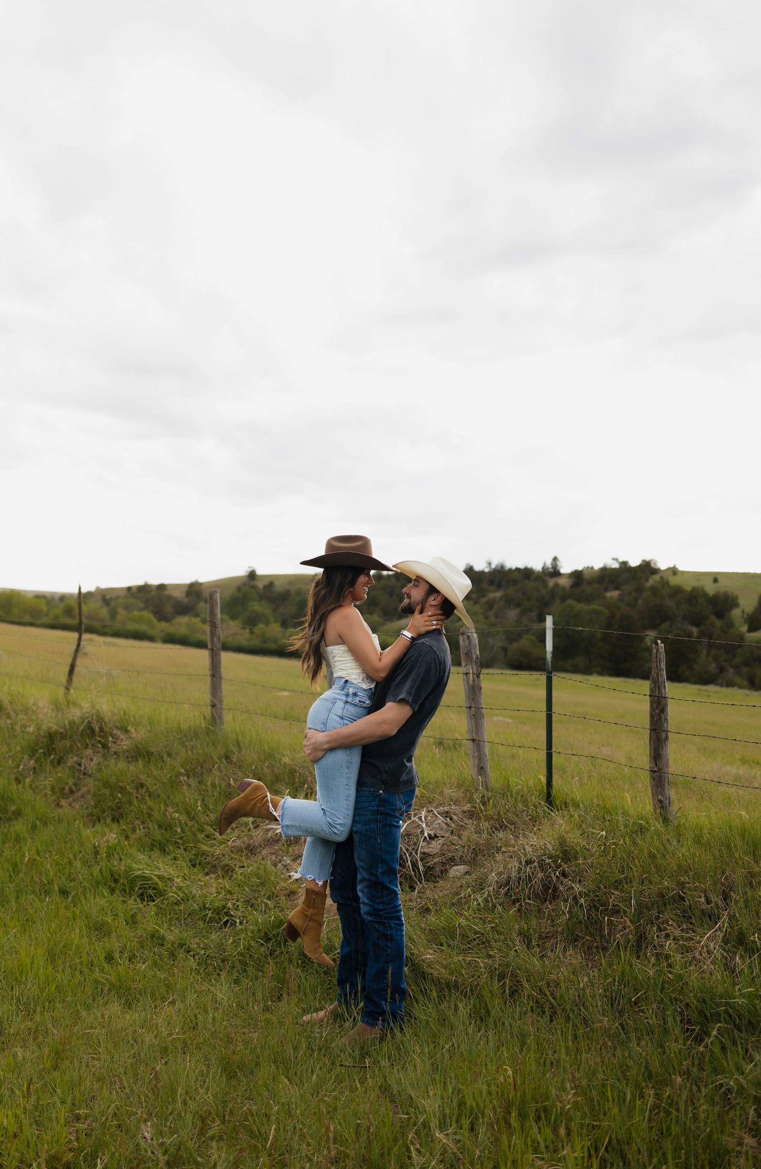 Couple in cowboy hats embracing in a green pasture, man lifting woman near a fence on an overcast day.