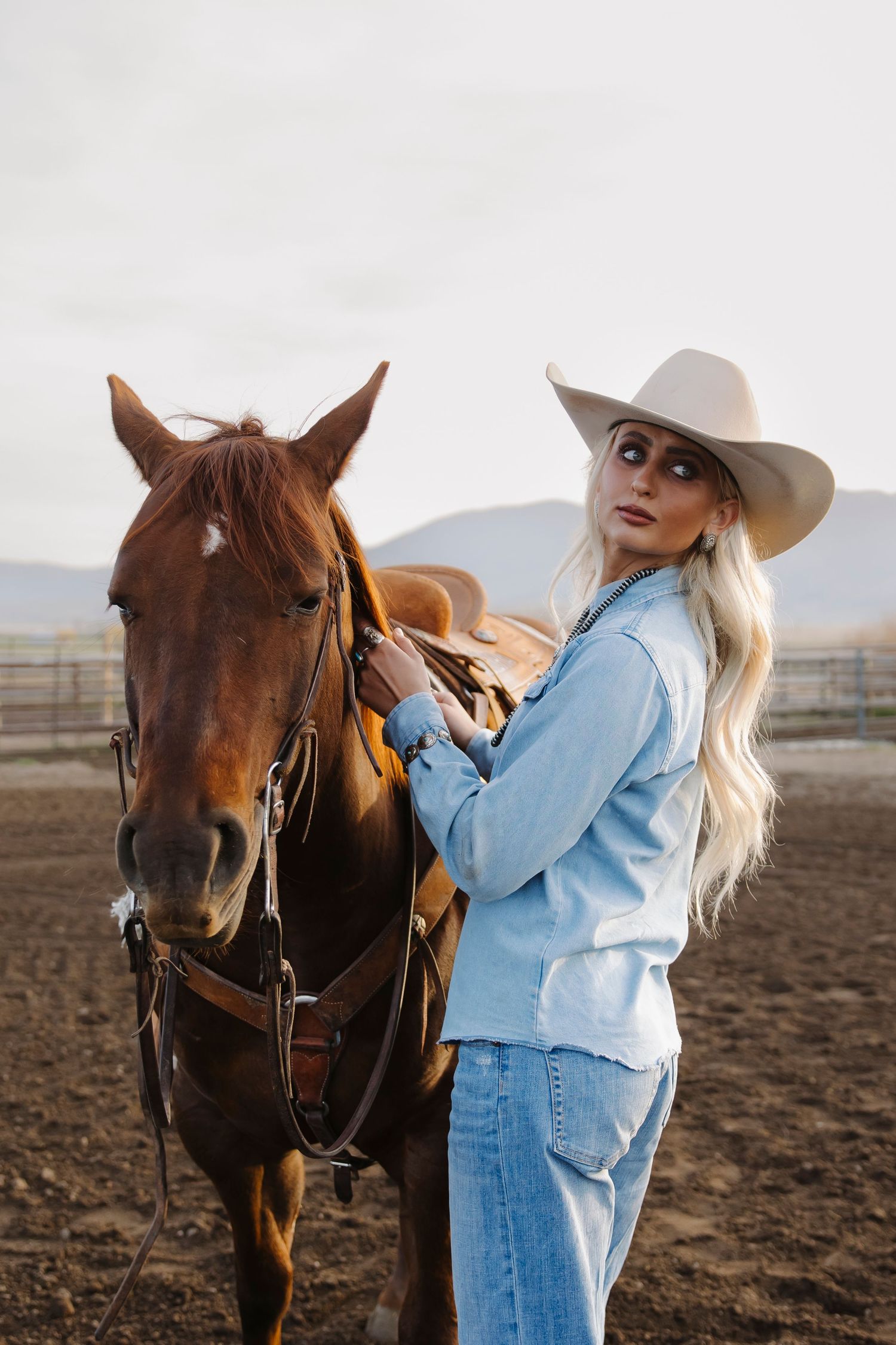 Blonde woman in white cowboy hat and jeans adjusting saddle on chestnut horse in outdoor ranch setting.