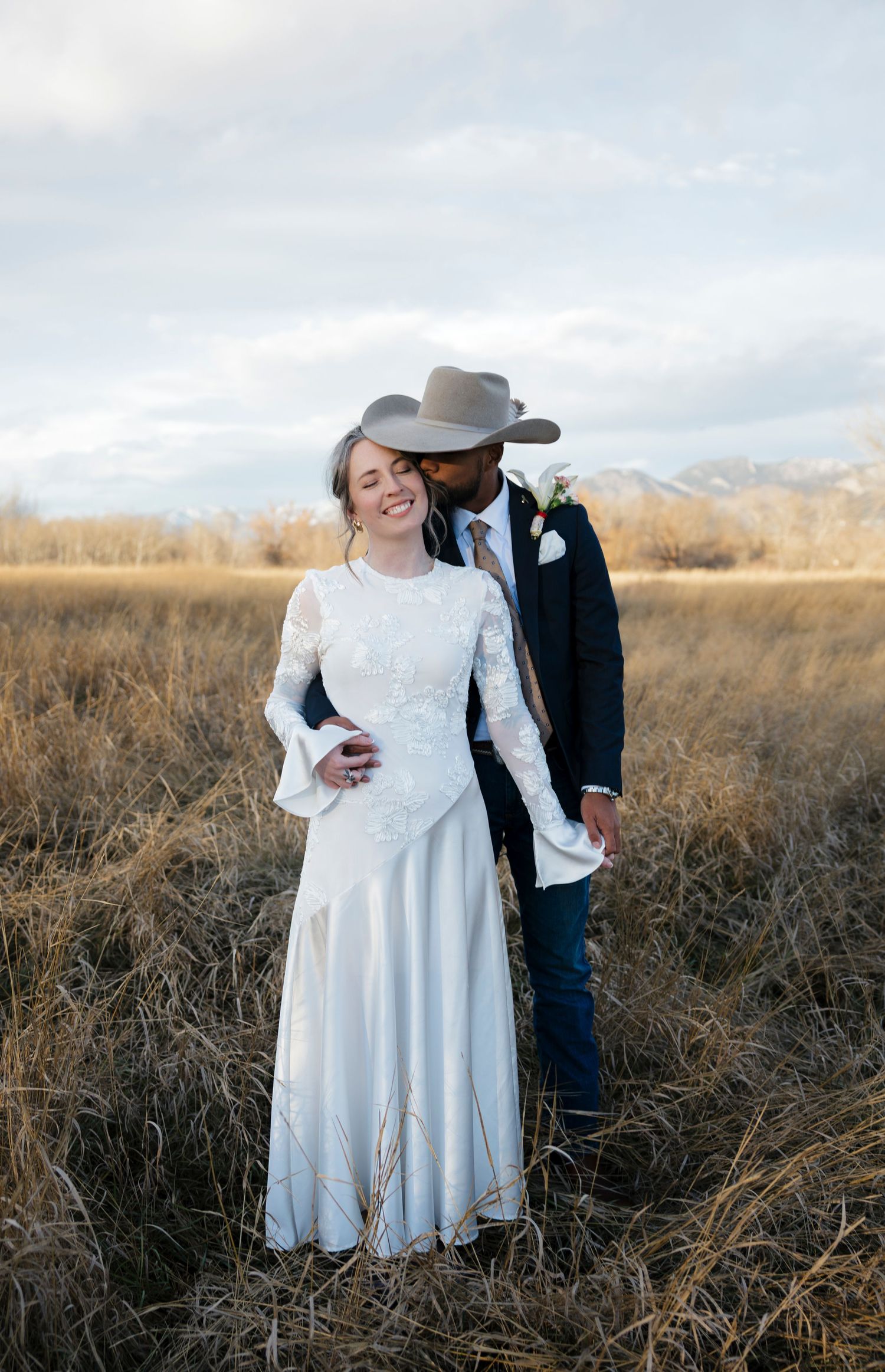 Intimate wedding portrait in rustic field with mountains and cloudy sky backdrop.