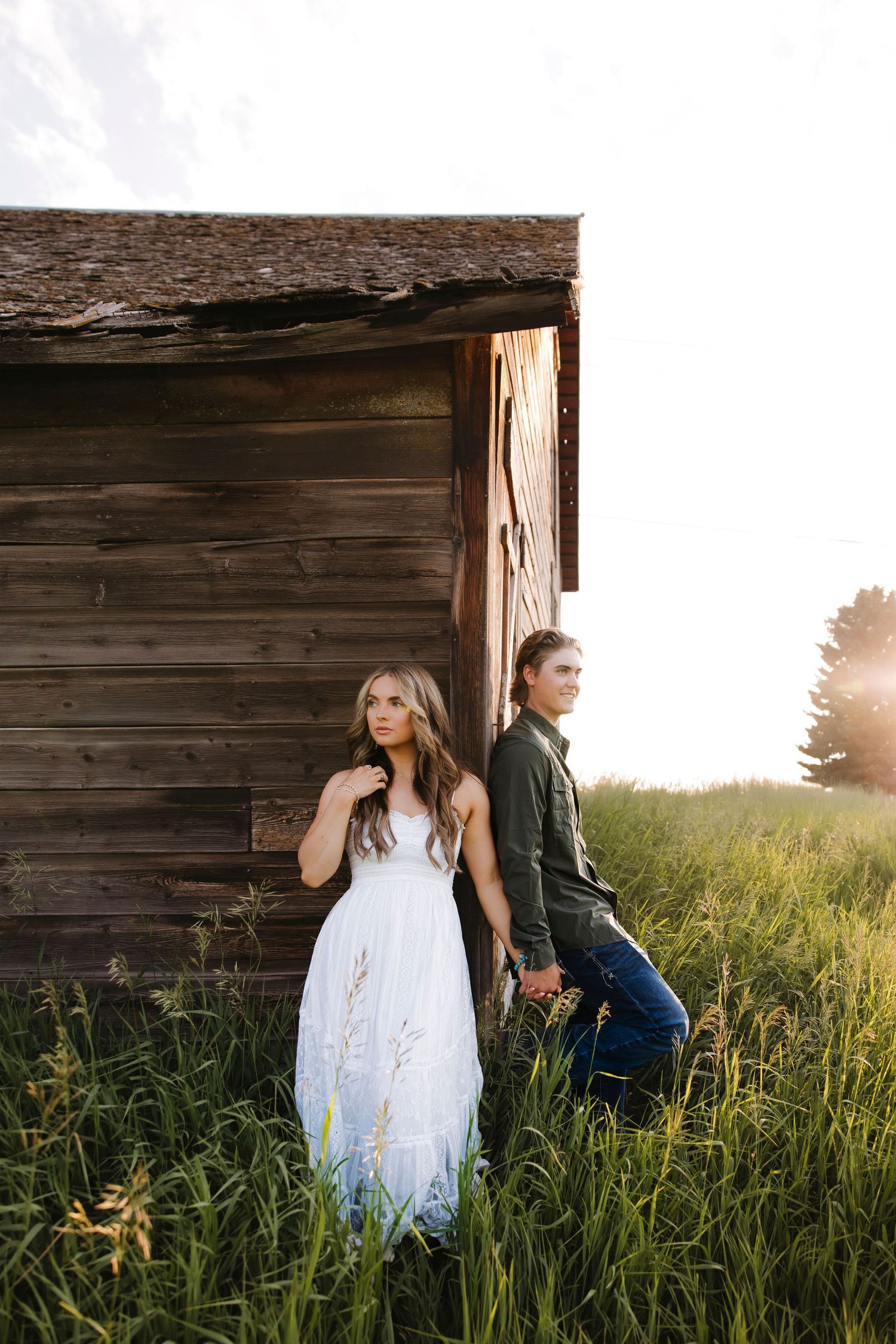 Couple posing against a rustic wooden barn wall surrounded by tall grass at golden hour.