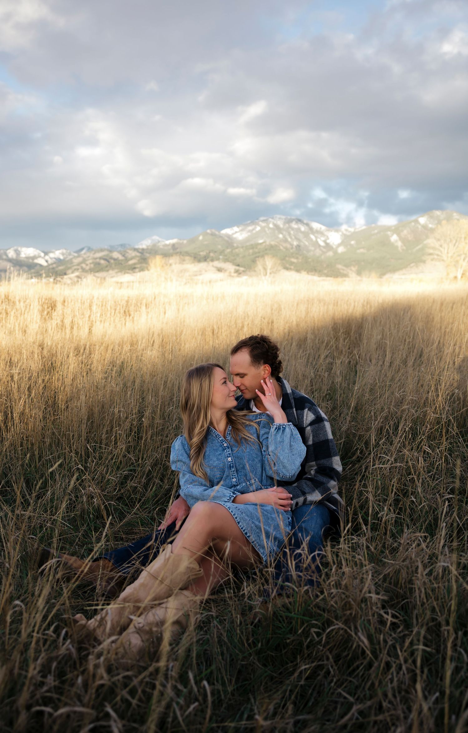 Romantic moment captured in tall grass with snow-capped mountains and cloudy skies.