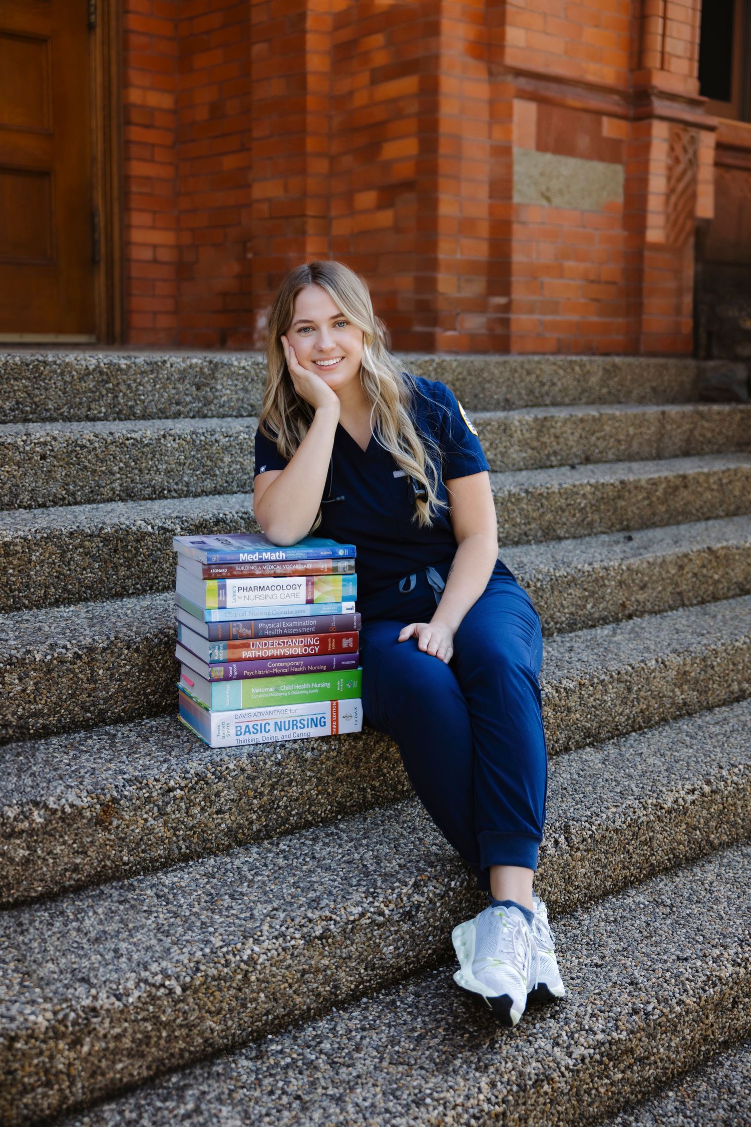 Nursing student in blue scrubs sits on steps with textbooks.