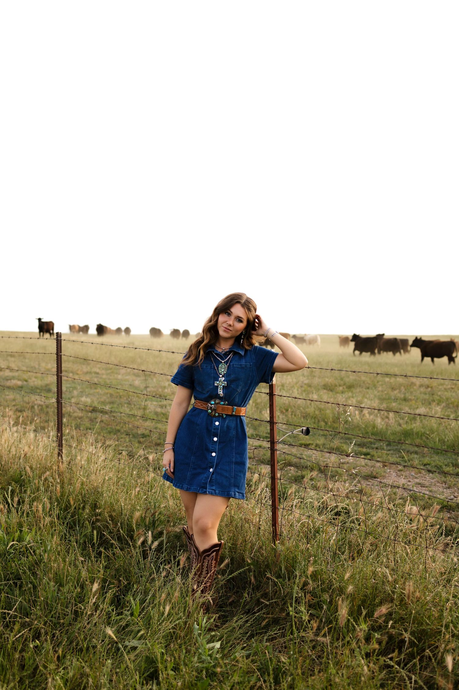 Young woman in denim dress and cowboy boots poses by barbed wire fence with cattle grazing in background.