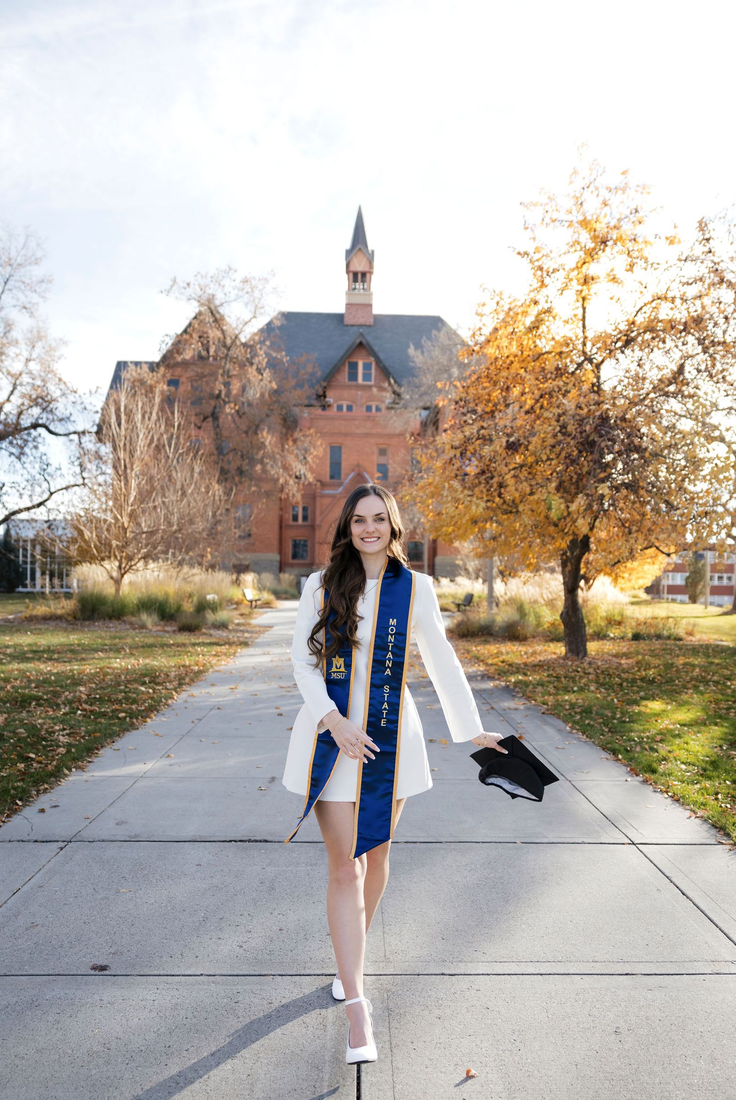 Graduate in white dress and blue stole poses on campus walkway during autumn.