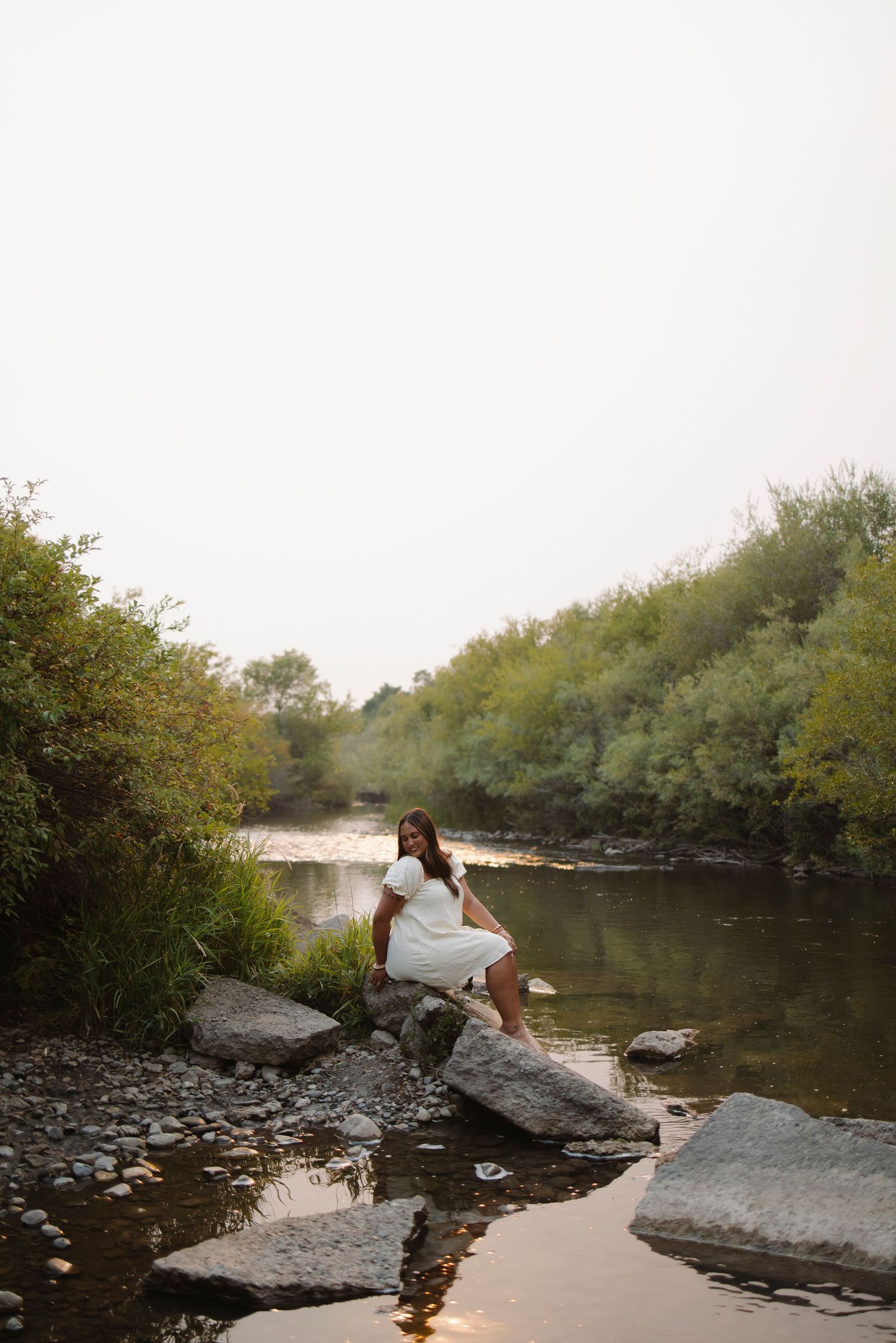 Woman in white dress sitting on rocks by a serene river surrounded by lush green trees.