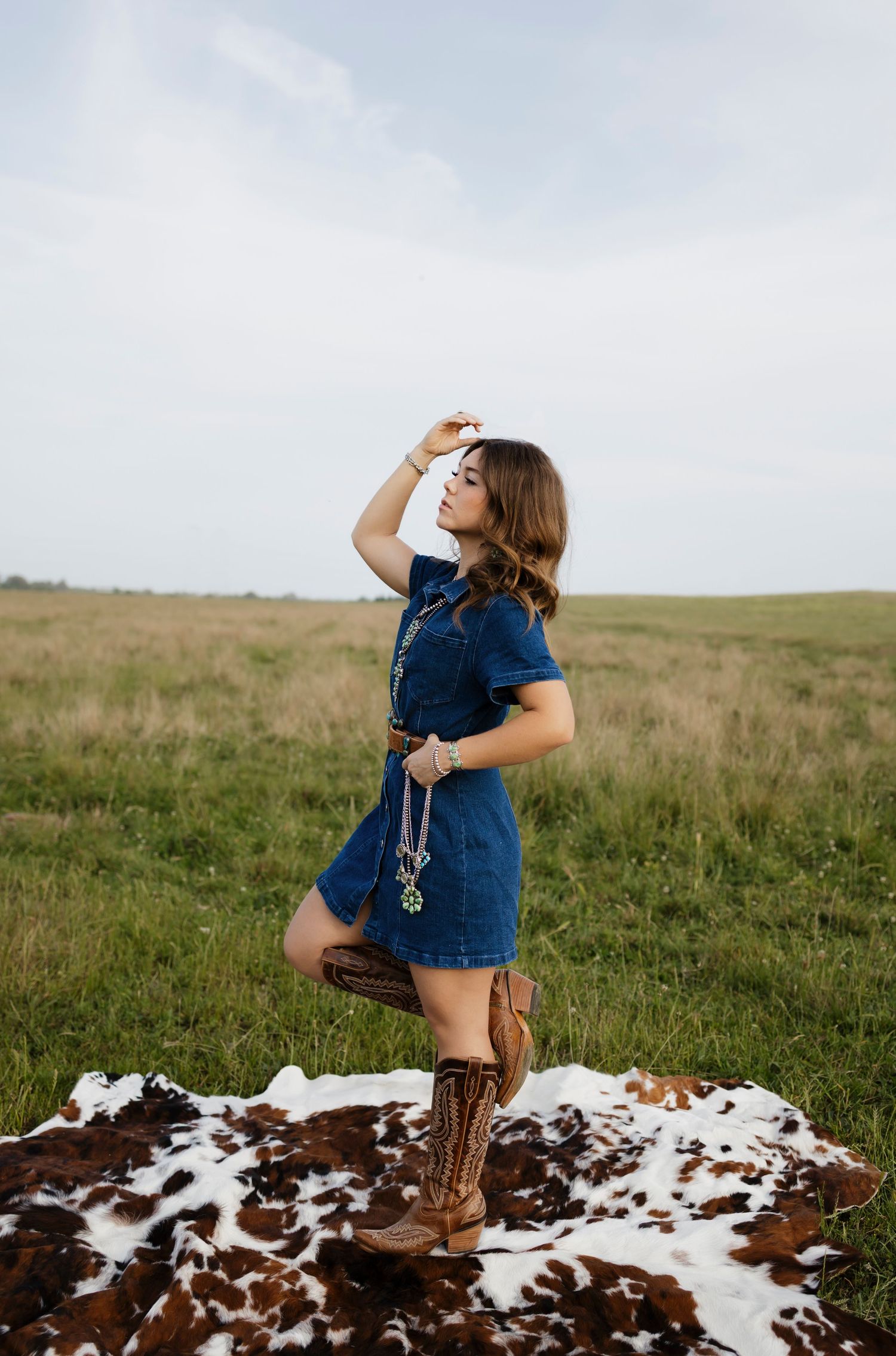 Young woman in denim dress and cowboy boots poses on cowhide rug in open green field.