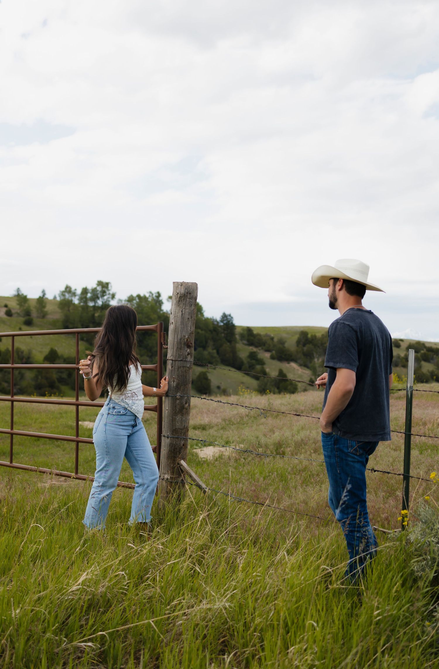 A man in a cowboy hat and woman in jeans stand by a rustic farm gate in a lush green rural landscape.