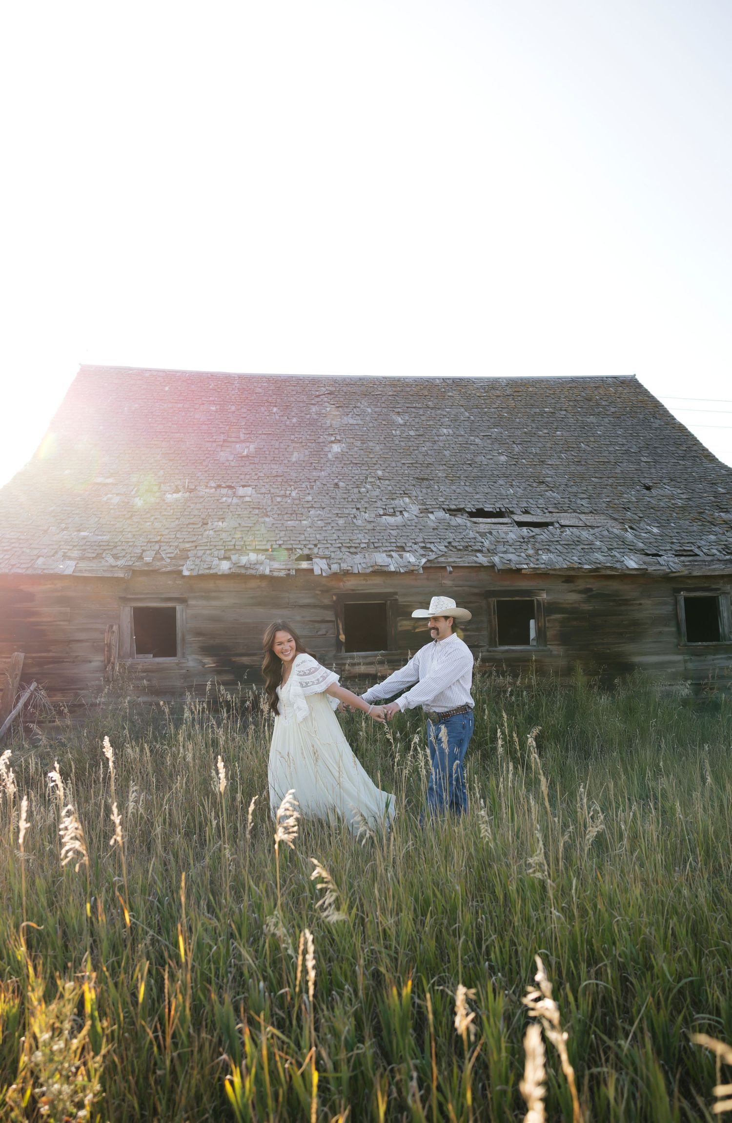 Couple dances in tall grass near old barn during golden hour photography session.