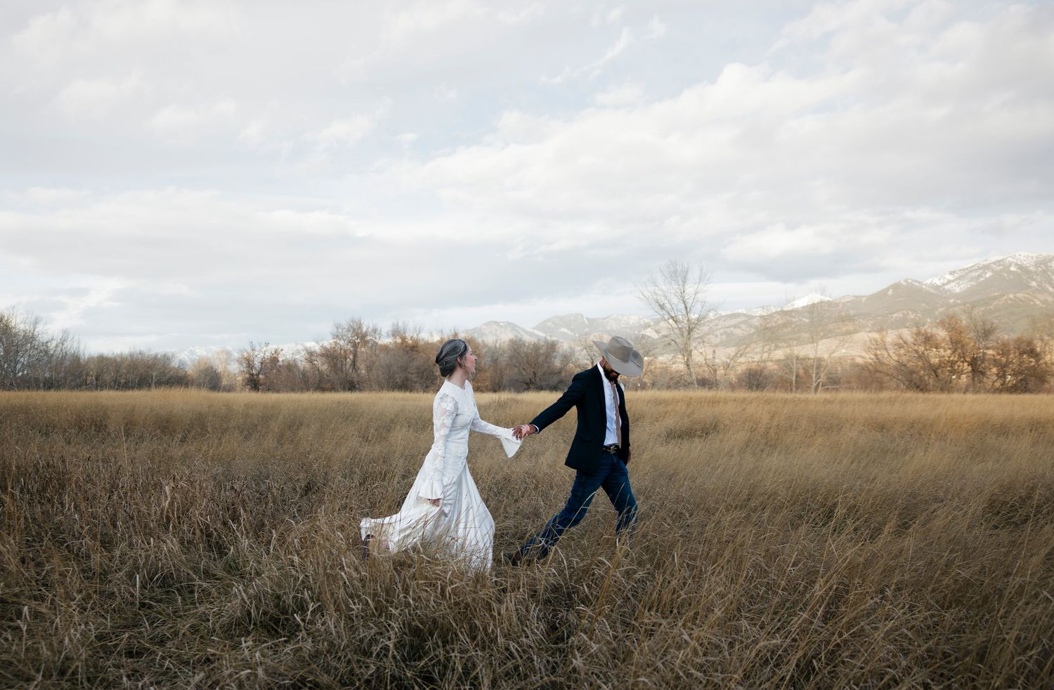 Couple walking hand in hand through golden field with cloudy sky overhead.